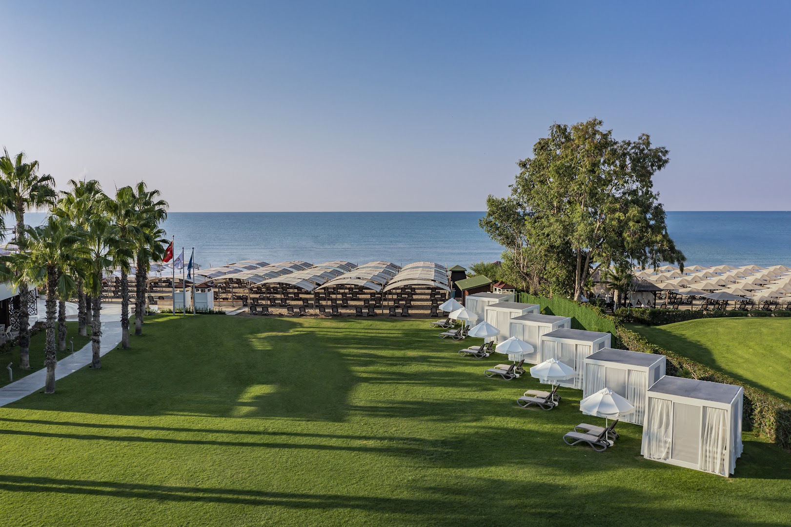 Papillon Zeugma Relaxury privātās pludmales zona ar zālāju, palmām un baltām atpūtas teltīm pie Vidusjūras