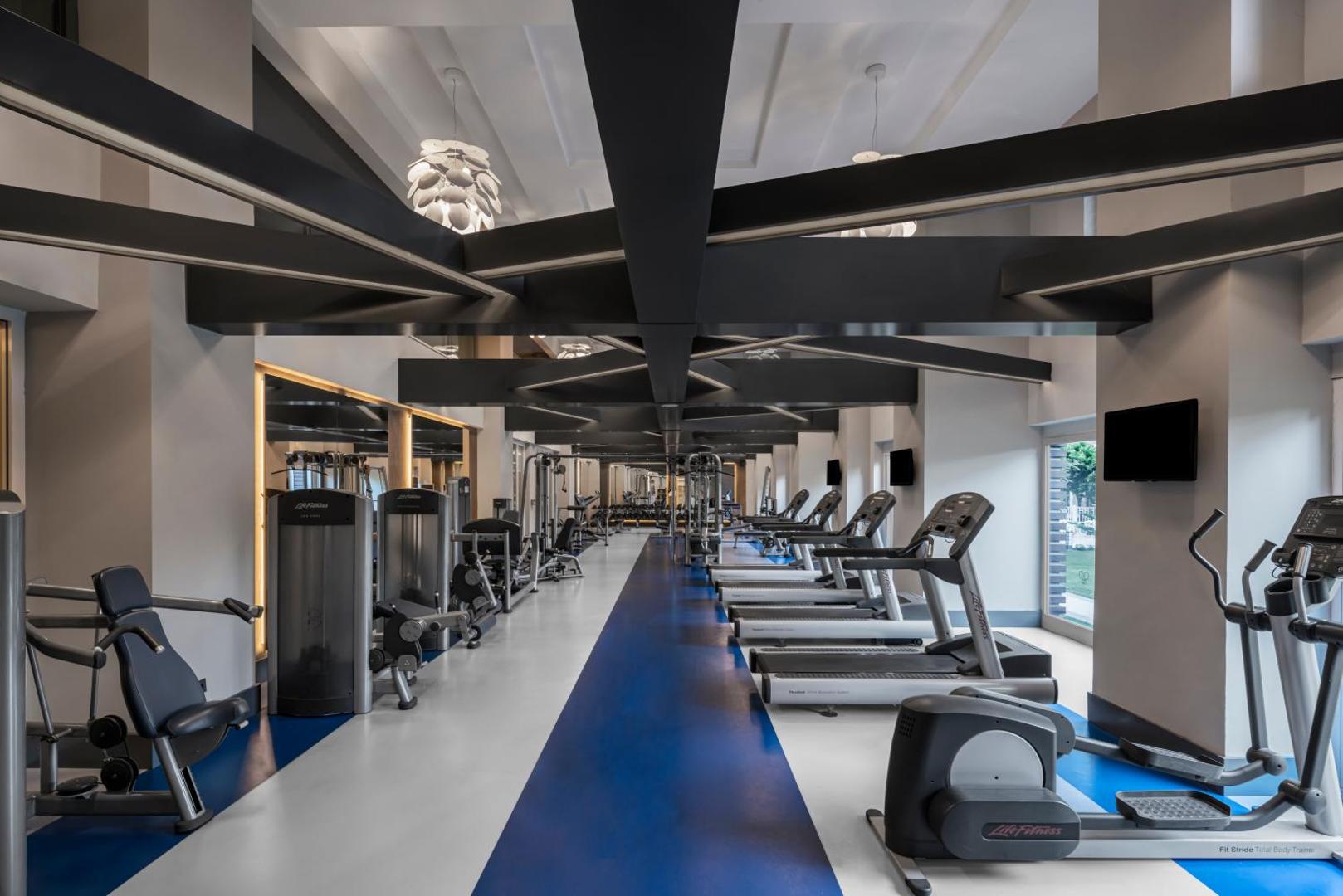 Papillon Ayscha mūsdienīgs fitness centrs ar Life Fitness skrejceļiem un trenažieriem gaišā zālē ar augstiem griestiem