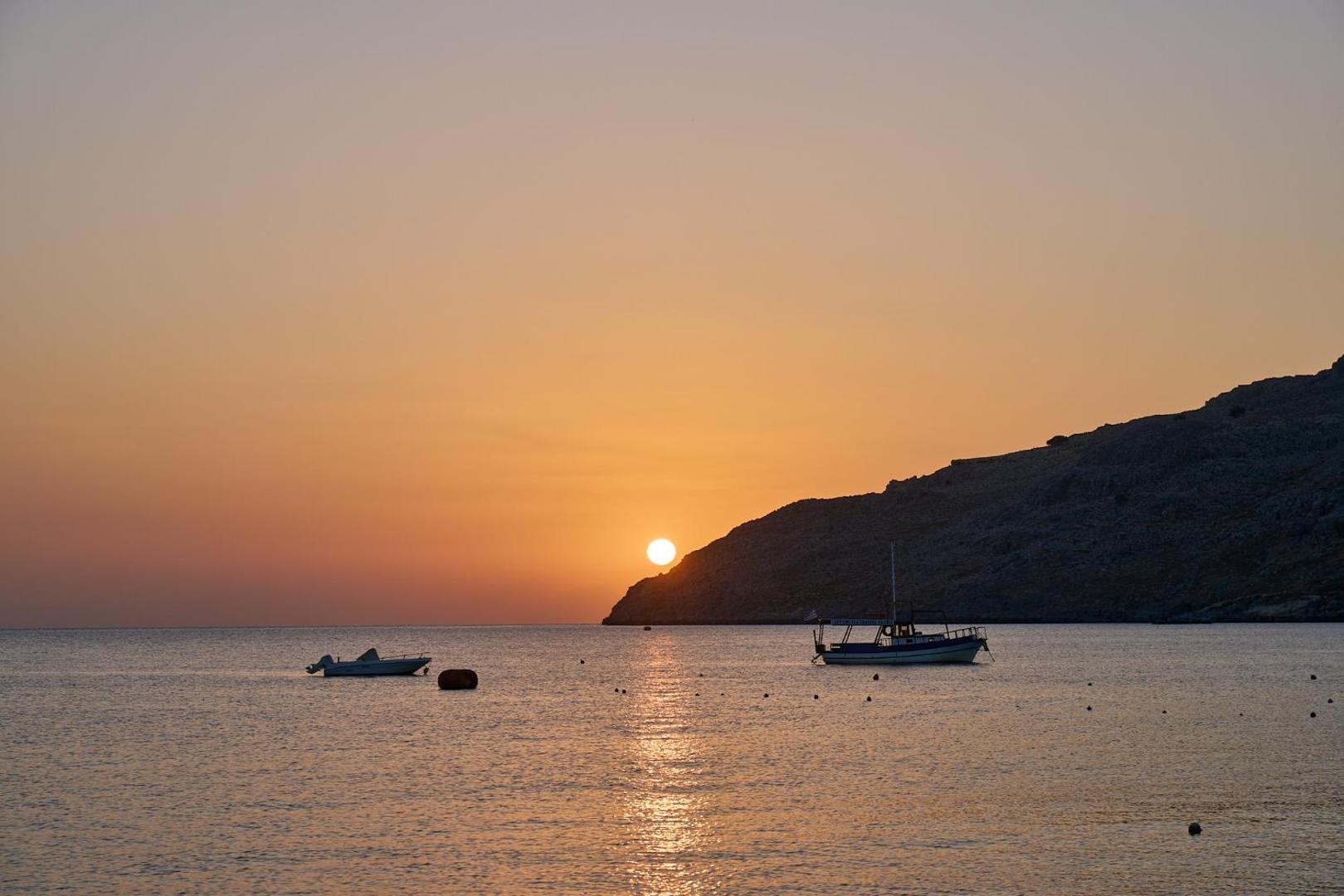 Lindos Royal Resort skats uz saulrietu Vidusjūrā Rodas salā — kalnu siluets un nelielas laivas mierīgā līča ūdenī