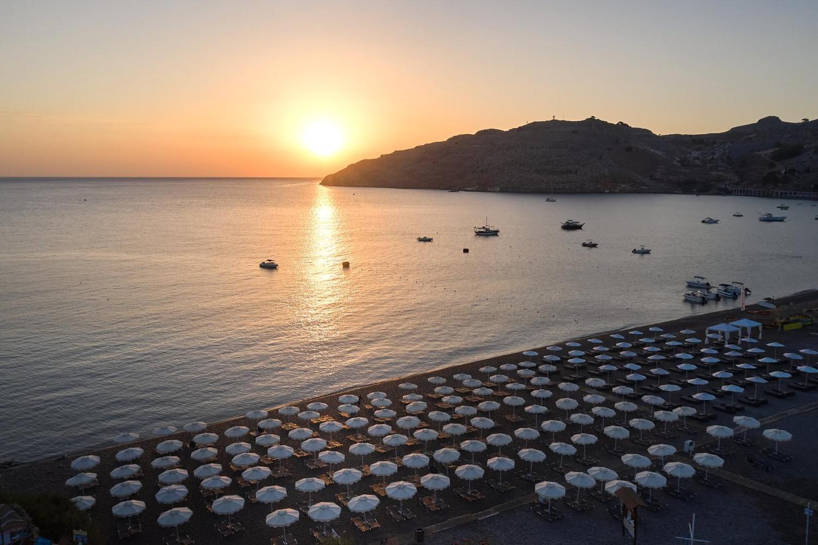 Saulriets virs Lindos Royal Resort līča Rodas salā — panorāmas skats uz jūru, kalniem, jahtām un pludmali vakara gaismā