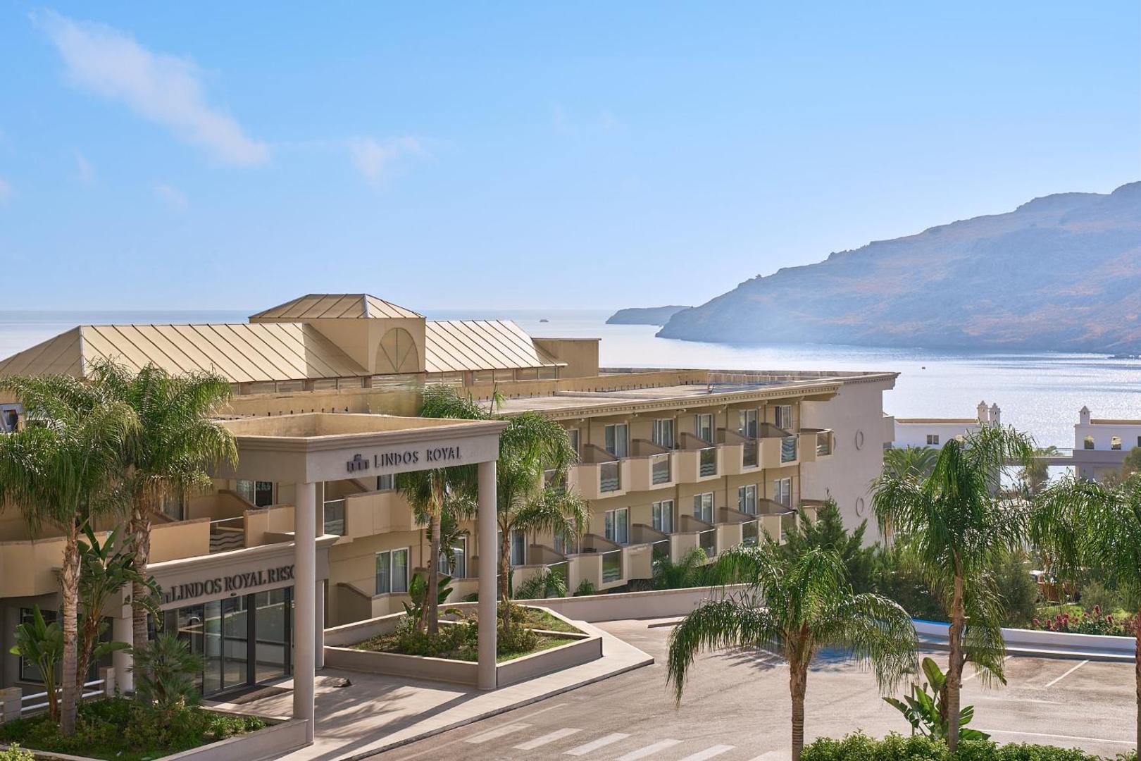 Lindos Royal Resort galvenā ieeja un fasāde ar palmām, fonā redzami Rodas salas kalni un jūra saulainā dienā