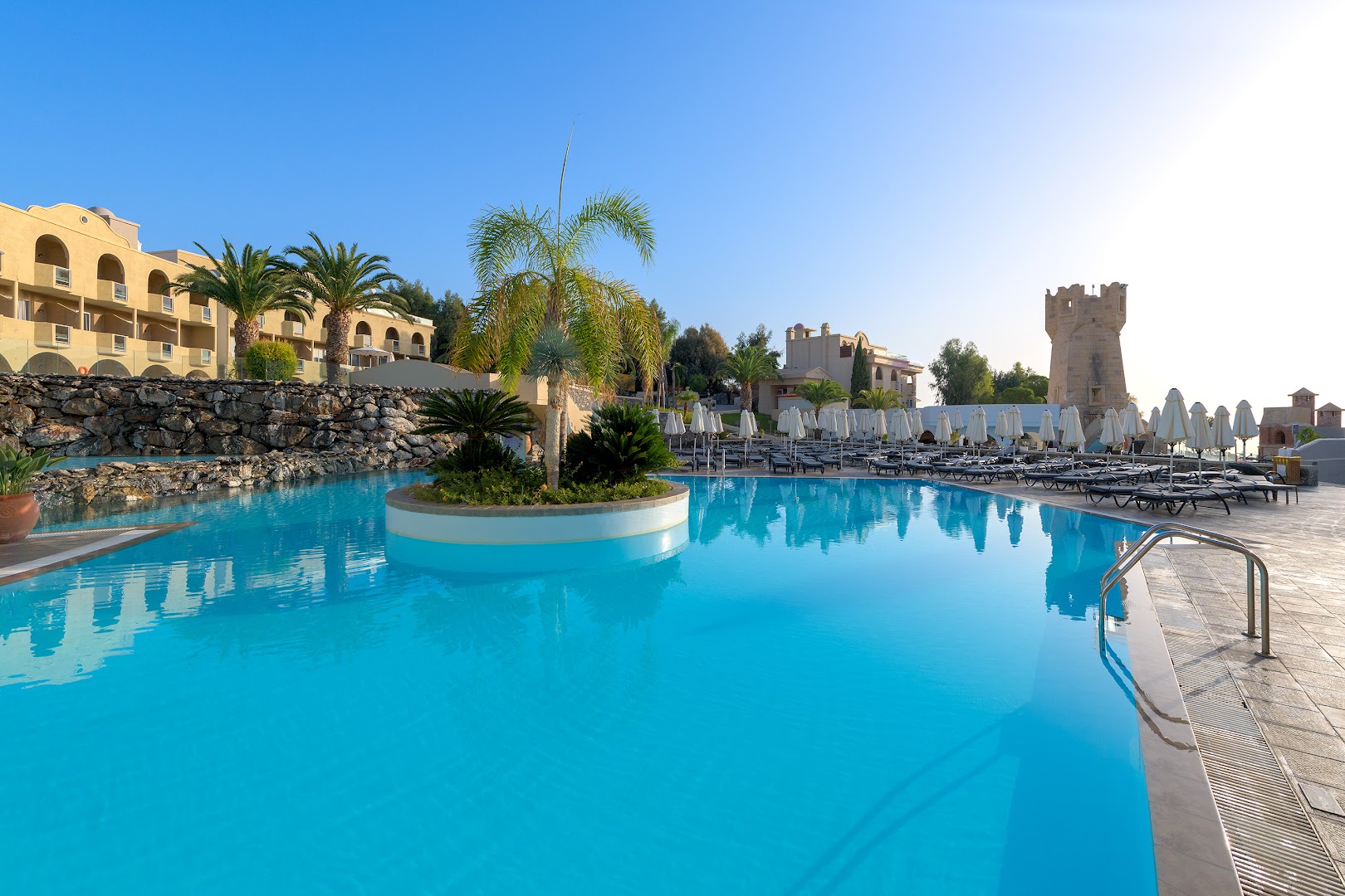 Lindos Royal Resort āra baseins ar dekoratīvu palmu salu un skatu uz viesnīcas korpusiem un akmens sienu