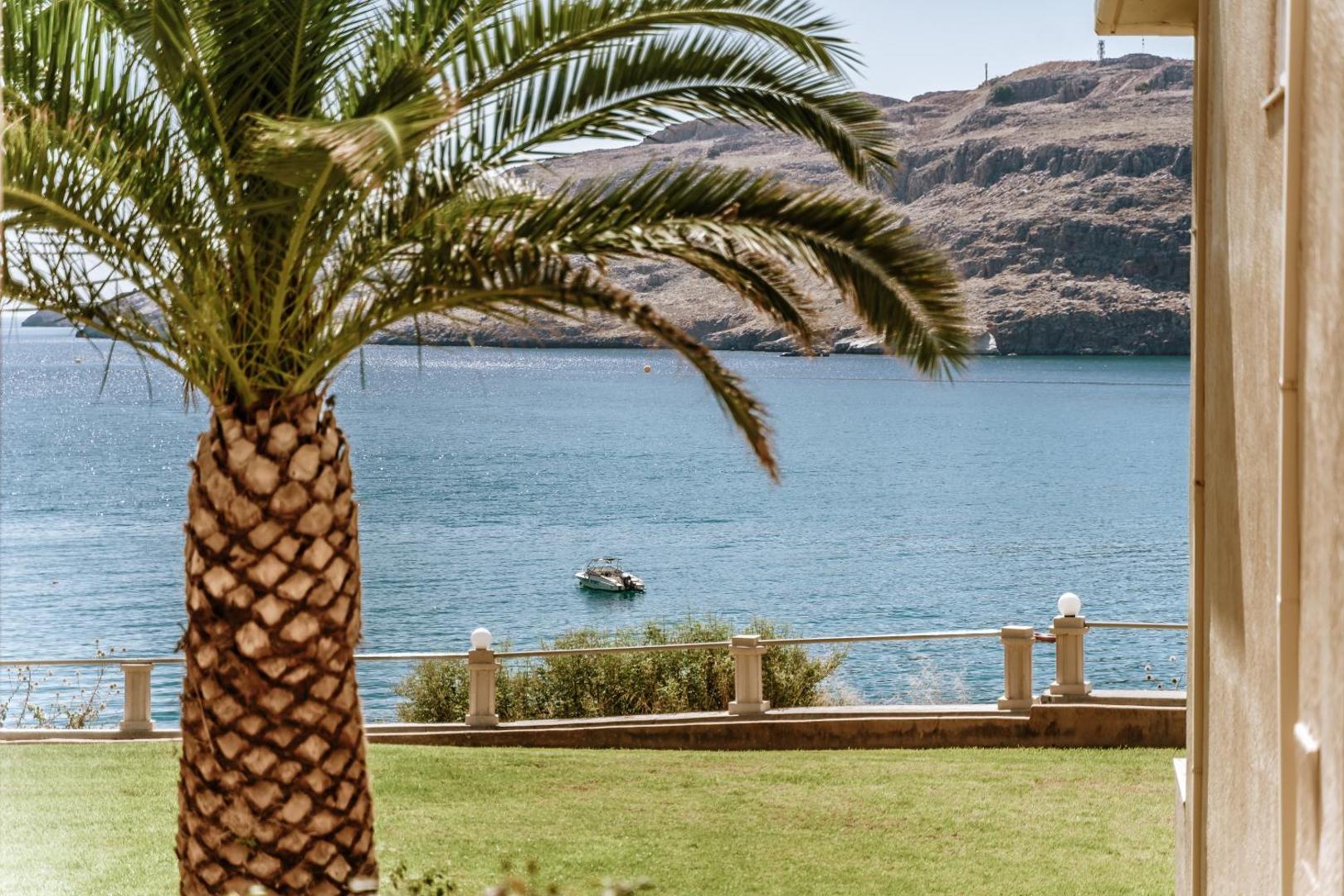 Lindos Royal Resort skats no viesnīcas dārza uz jūru Rodā — palma priekšplānā un zils līcis ar kalniem fonā