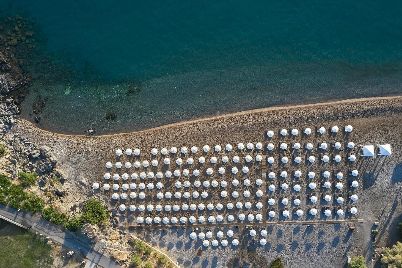 Lindos Royal Resort privātā pludmale no putna lidojuma — dzidrs jūras ūdens, smilšu un oļu krasts ar simtiem baltu saulessargu