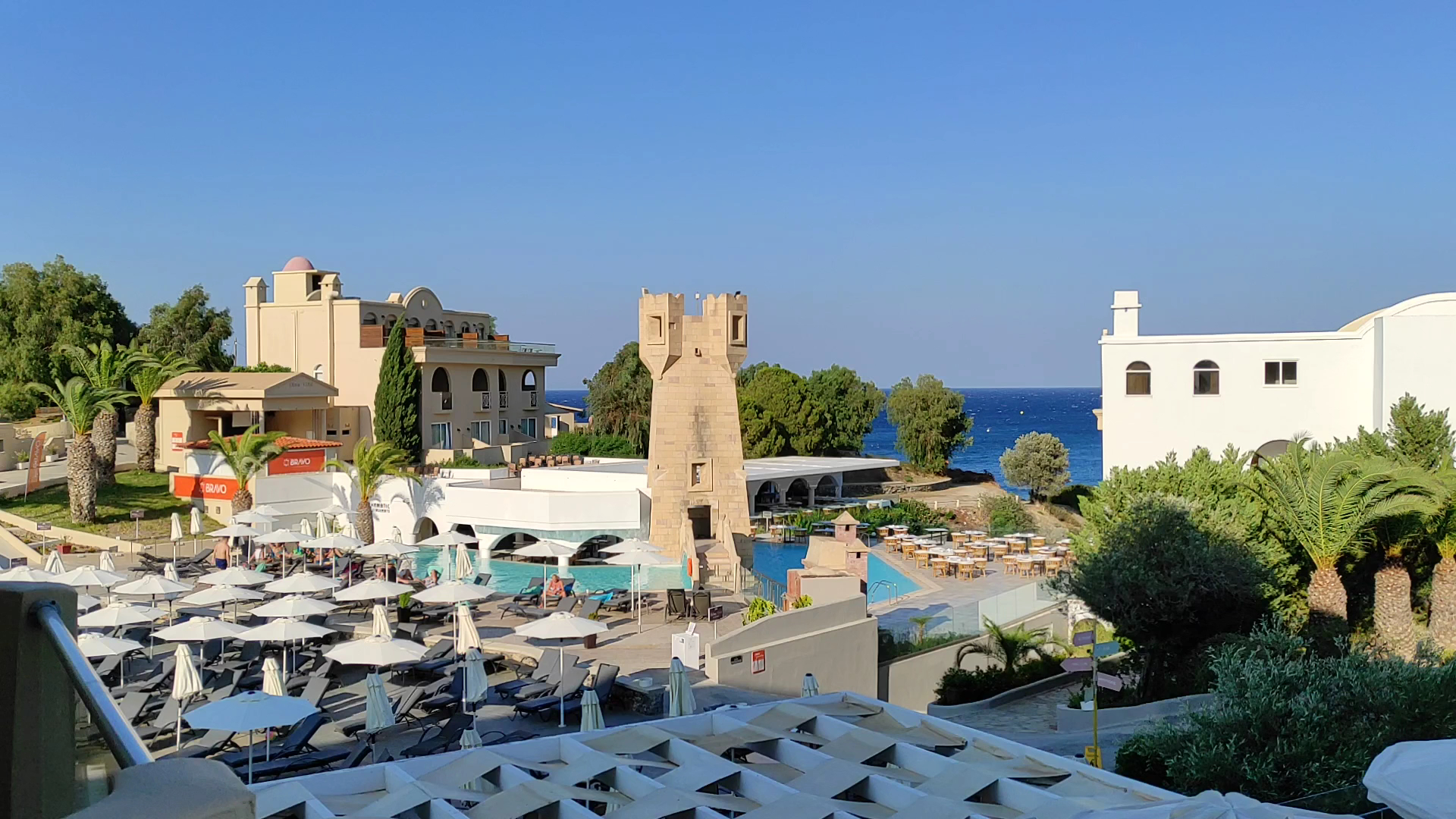 Lindos Royal Resort baseinu zona ar dekoratīvu torni, saulessargiem un atpūtas krēsliem, fonā jūra un zilas debesis
