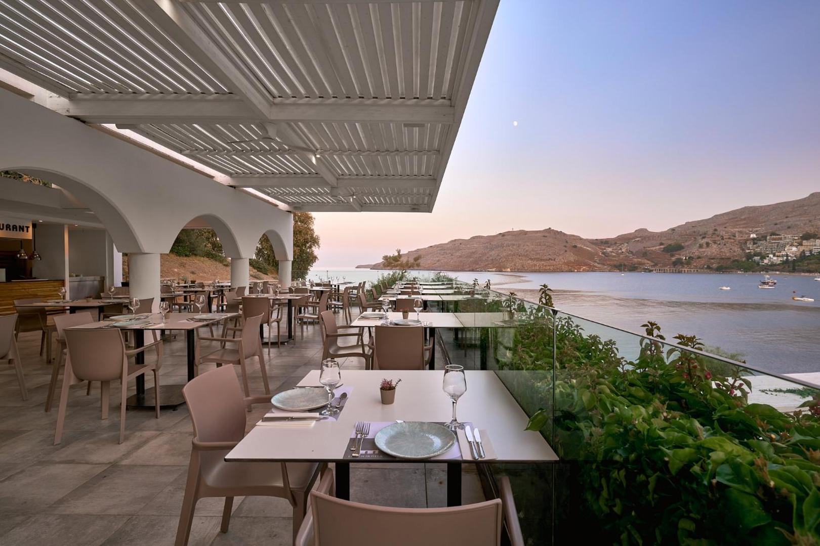 Lindos Royal Resort āra terases restorāns krēslas stundā — klāti galdi pie stikla margām ar skatu uz līci Rodā