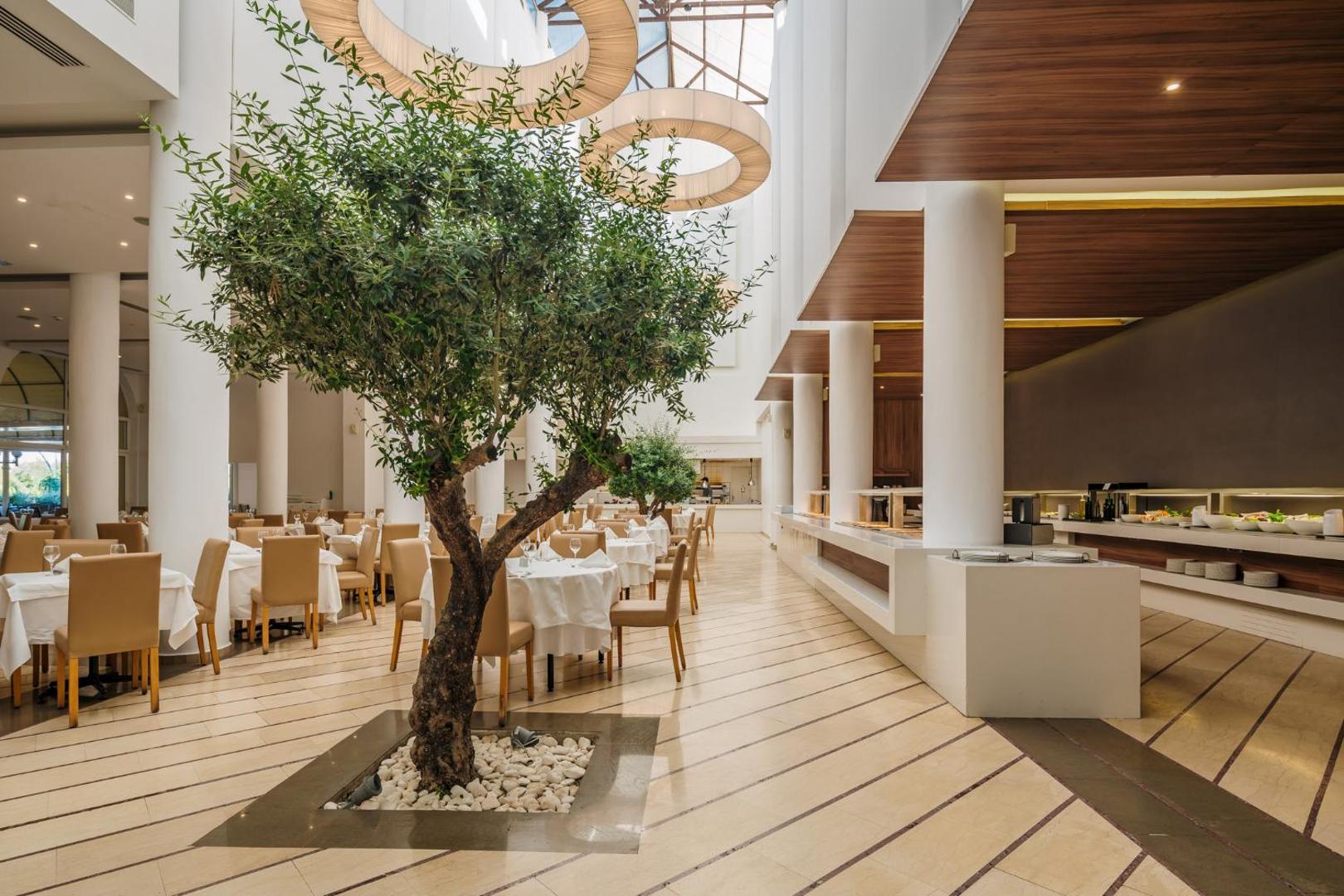Lindos Royal Resort galvenais restorāns ar olīvkoku centrā — moderna bufetes tipa zāle ar augstiem griestiem Rodā