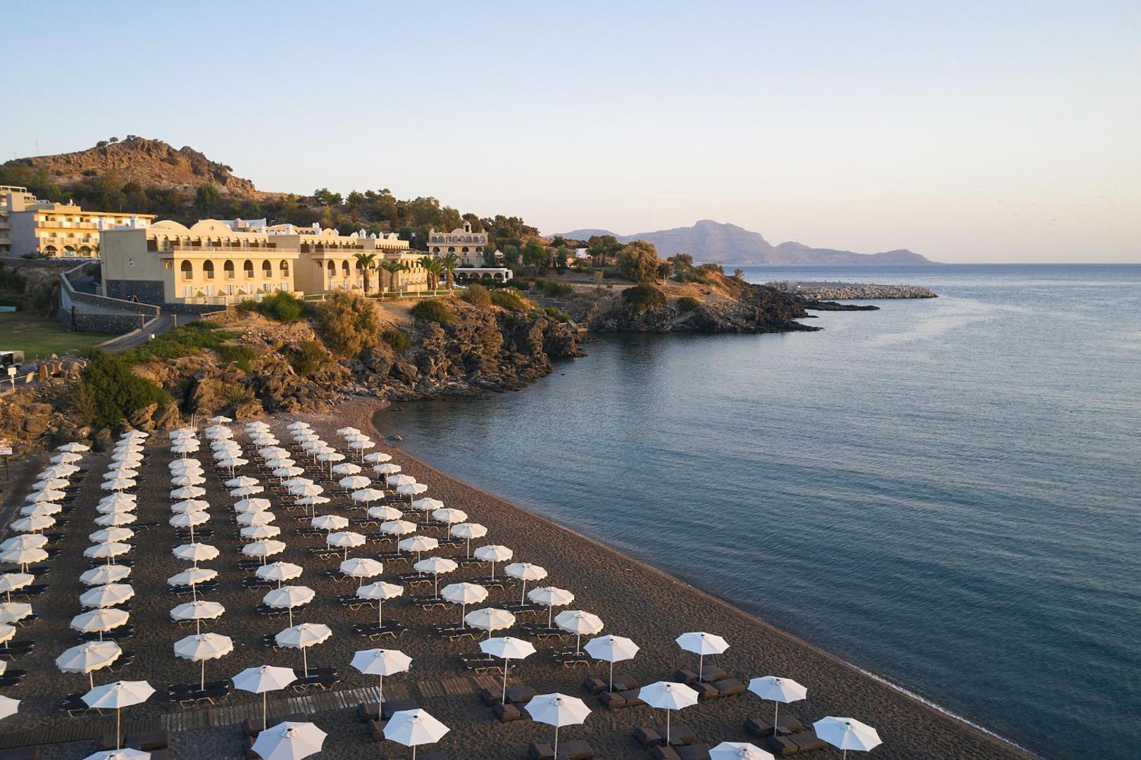 Lindos Royal Resort pludmales zona ar daudziem baltiem saulessargiem un guļamkrēsliem vakarā, fonā viesnīcas ēkas uz klints