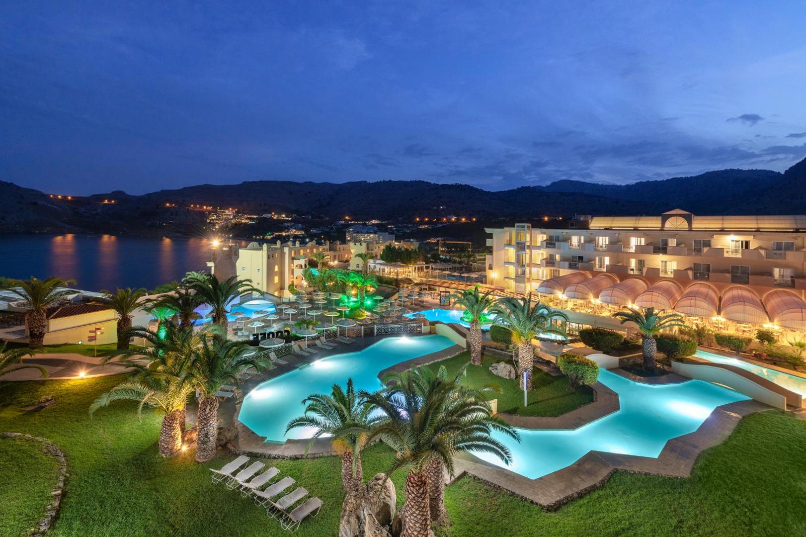 Lindos Royal Resort viesnīcas teritorija vakarā ar izgaismotiem baseiniem un ēkām uz kalnu un jūras līča fona Rodā