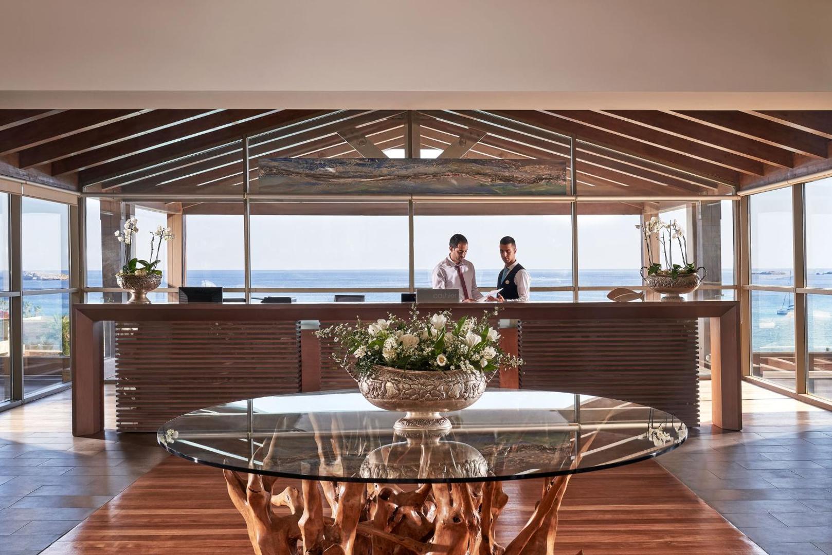 Viesnīcas Aquagrand Exclusive Deluxe Resort recepcijas zona ar koka apdari, stikla galdu un skatu uz zilo jūru fonā