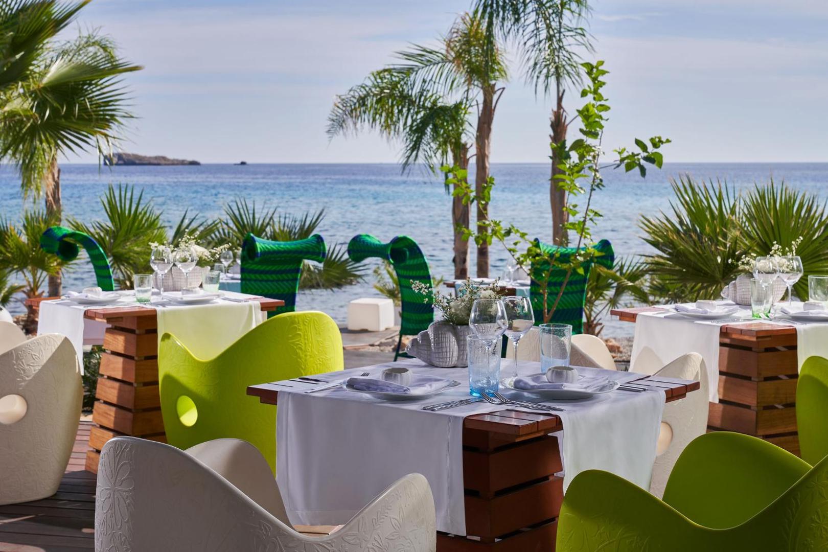 Aquagrand Exclusive Deluxe Resort āra terases restorāns ar skatu uz jūru Rodā — balti klāti galdi un koši krēsli palmu ielokā