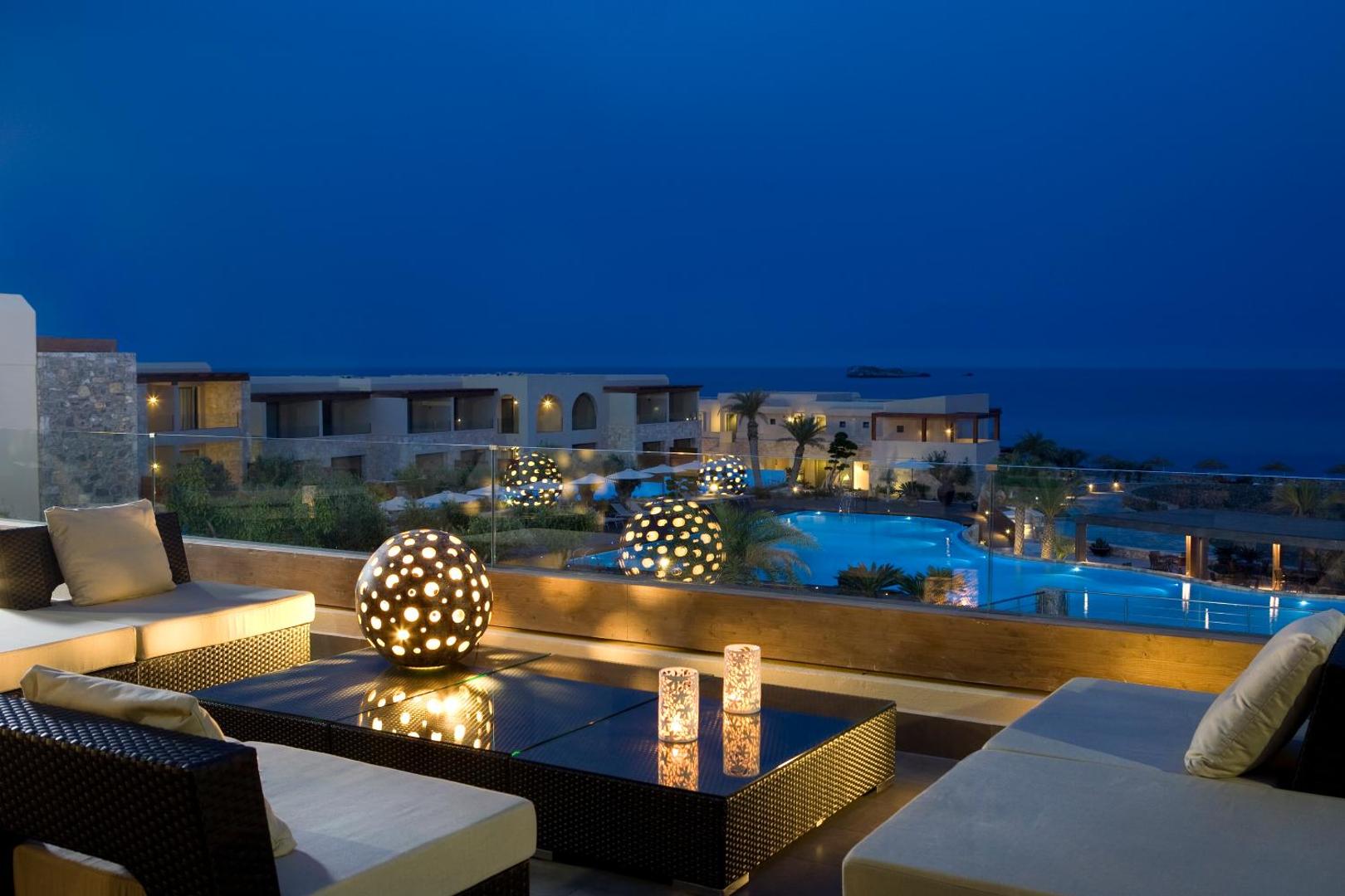 Aquagrand Exclusive Deluxe Resort terase vakarā ar skatu uz izgaismotu baseinu un jūru Rodas salā, Grieķijā — mīkstās mēbeles un dekoratīvās gaismas lodes