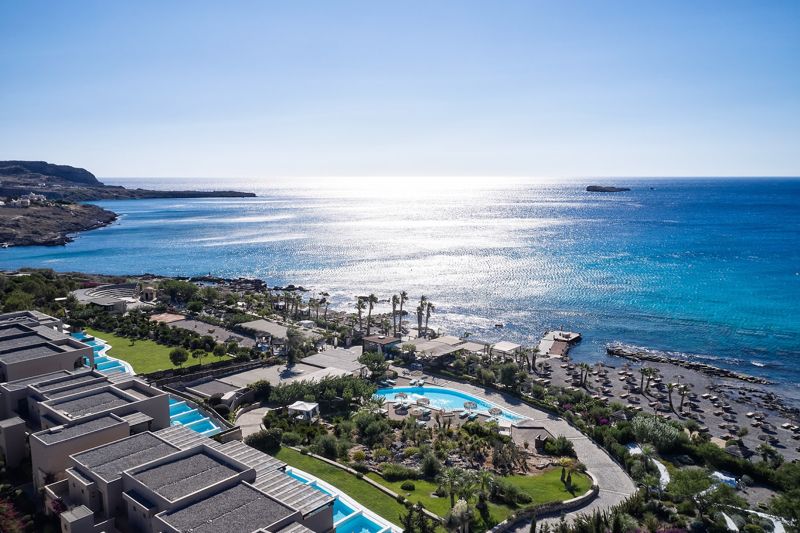 Aquagrand Exclusive Deluxe Resort dārzs ar ziedošiem augiem, palmām un viesnīcas korpusiem uz kalnu fona Rodas salā
