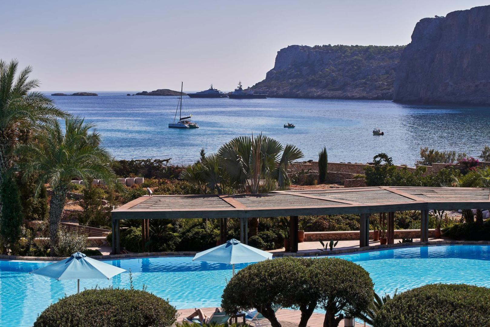 Aquagrand Exclusive Deluxe Resort skats no baseina zonas uz jūras līci ar jahtām un klintīm — Rodas salas piekrastes ainava