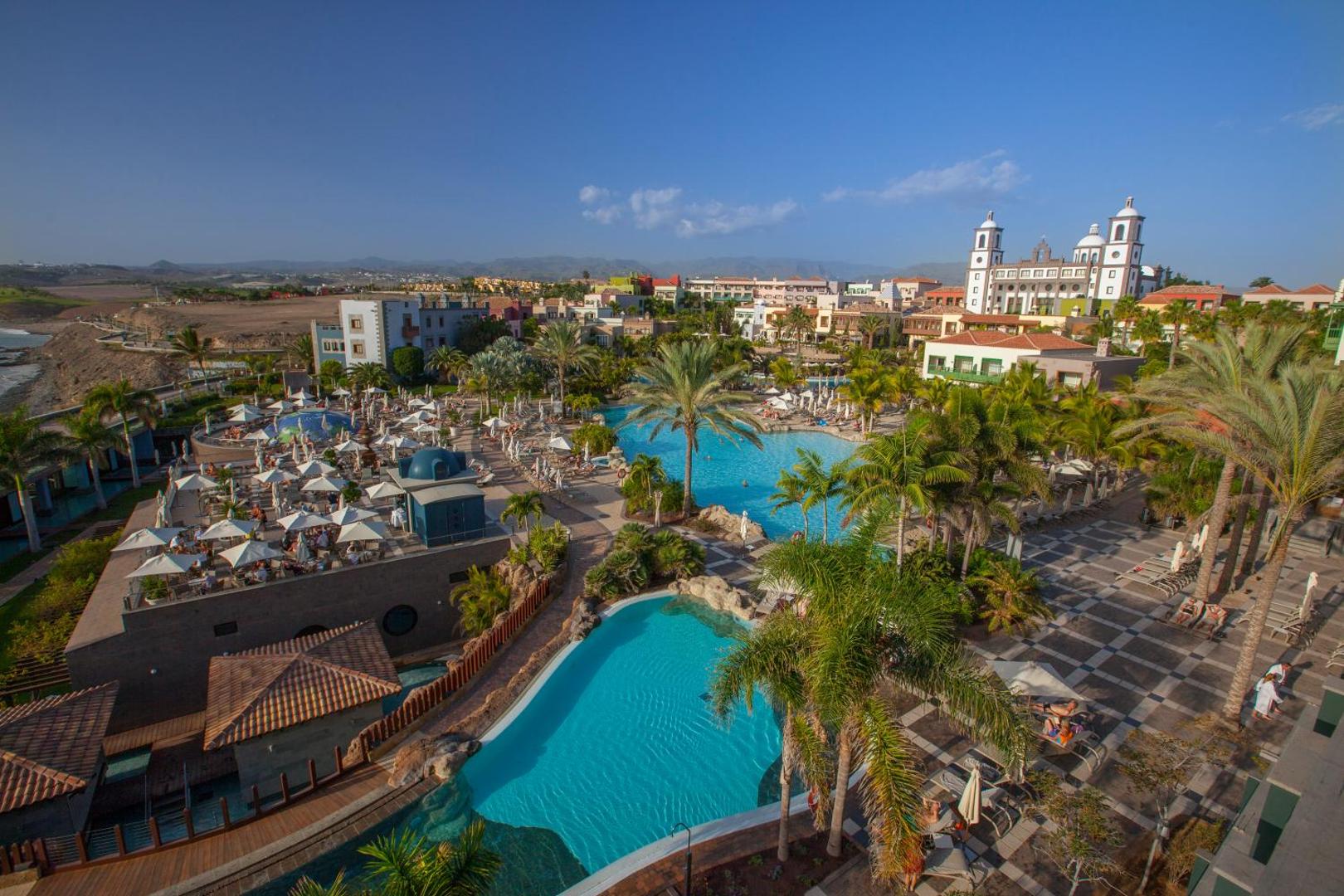 Lopesan Villa del Conde Resort & Thalasso plašā baseinu zona ar palmām un sauļošanās krēsliem — fonā redzama viesnīcas galvenā ēka