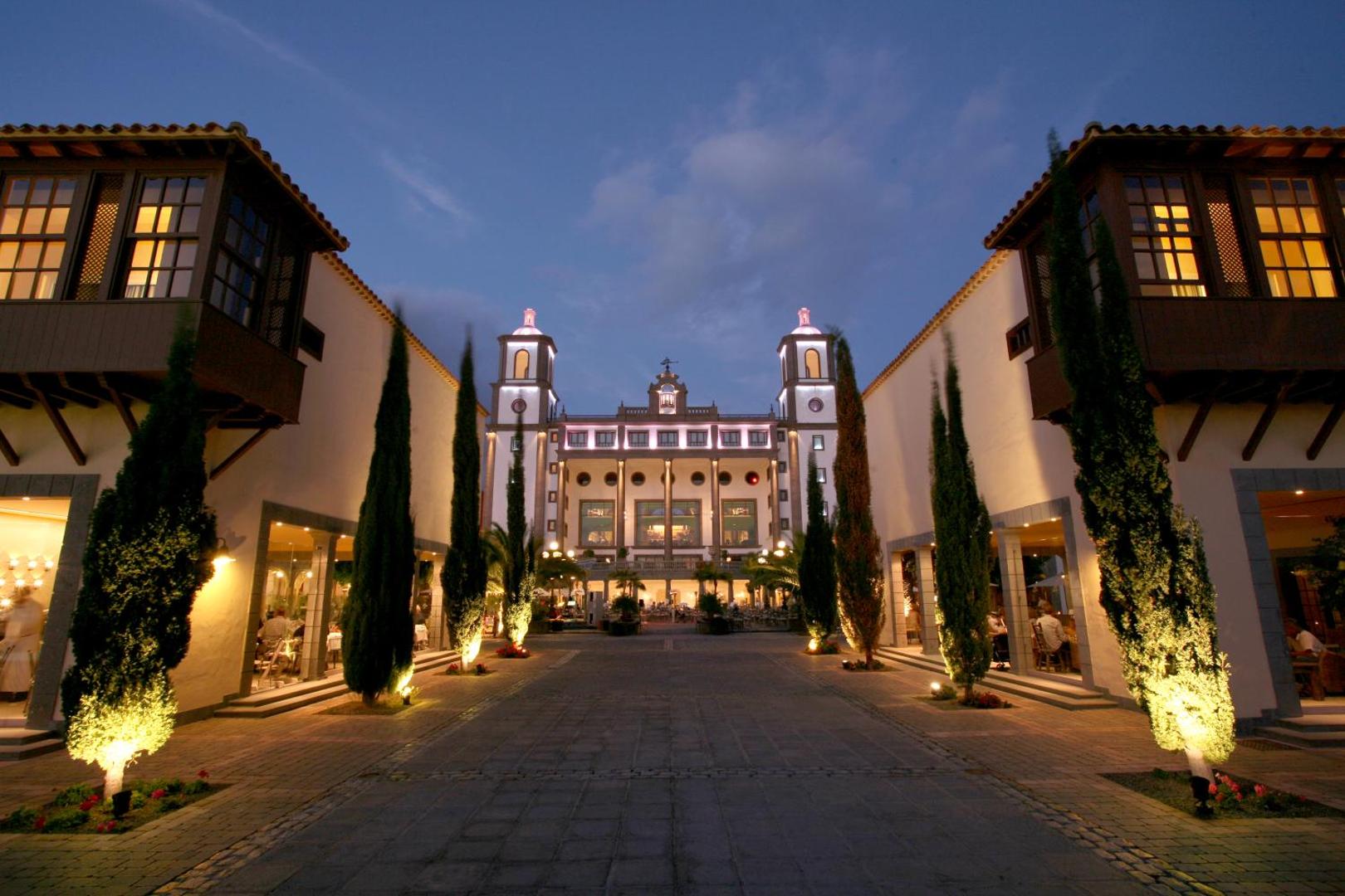 Lopesan Villa del Conde Resort & Thalasso centrālais laukums vakarā ar izgaismotu fasādi un ciprešu alejām — eleganta arhitektūra