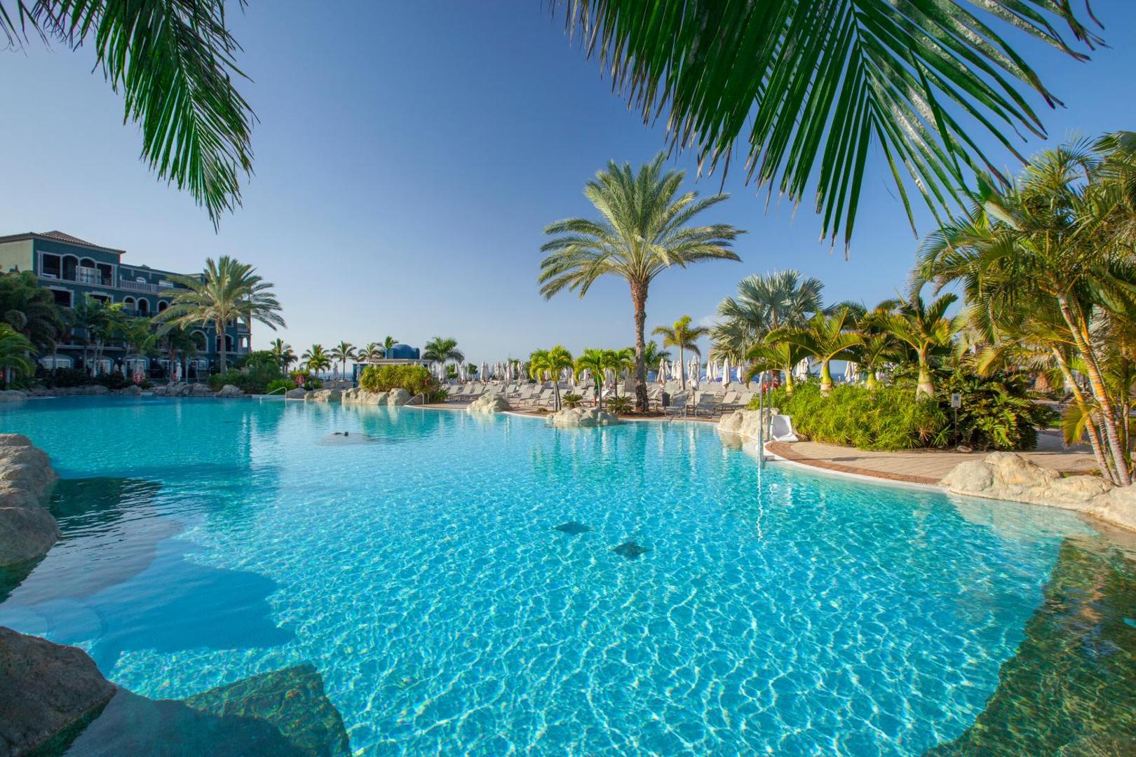 Lopesan Villa del Conde lagūnas tipa baseins ar kristāldzidru ūdeni, palmām un klintīm saulainā dienā