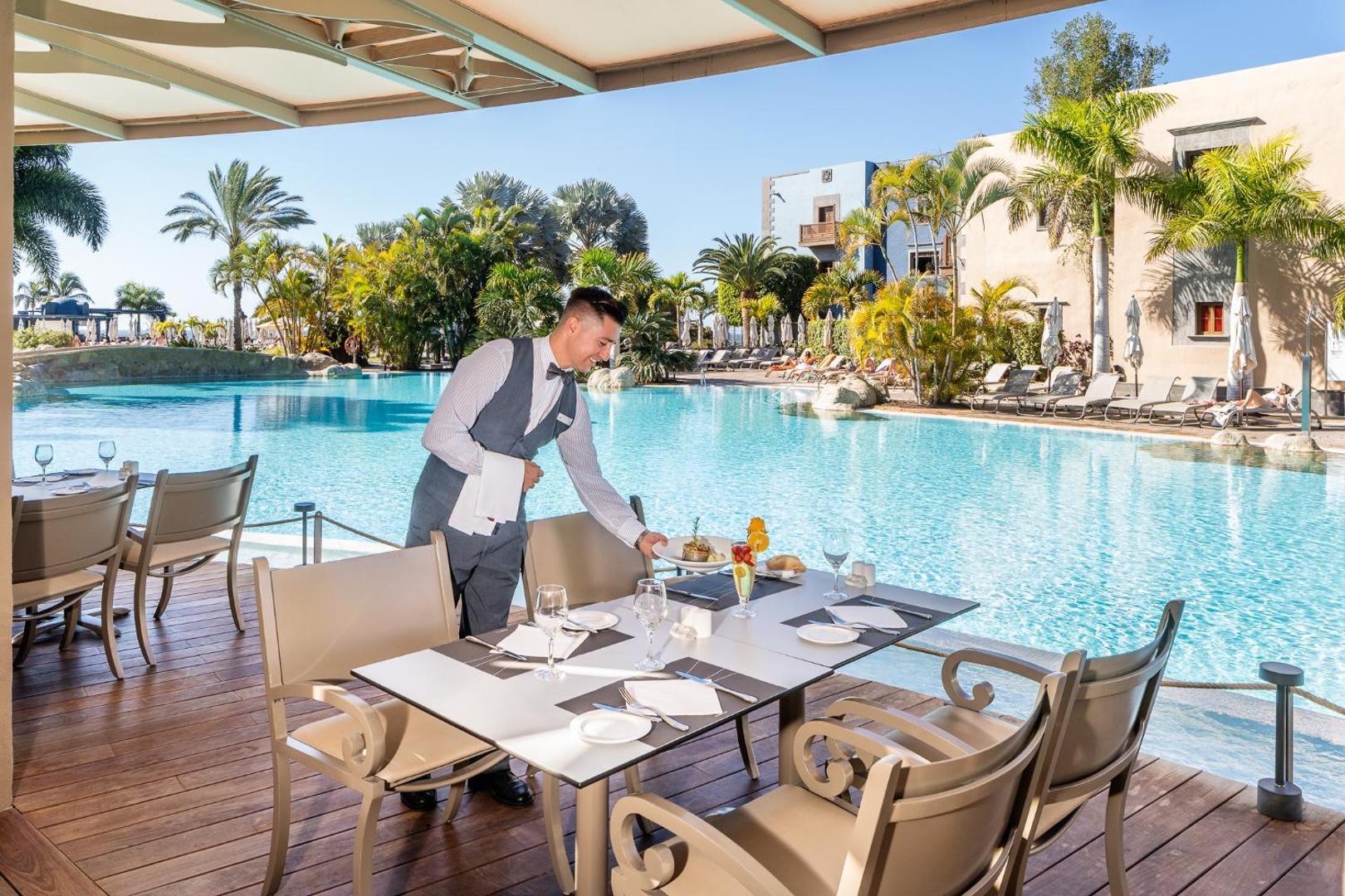 Viesmīlis klāj galdu Lopesan Villa del Conde Resort & Thalasso āra terasē pie baseina, fonā palmas un kūrorta ēkas