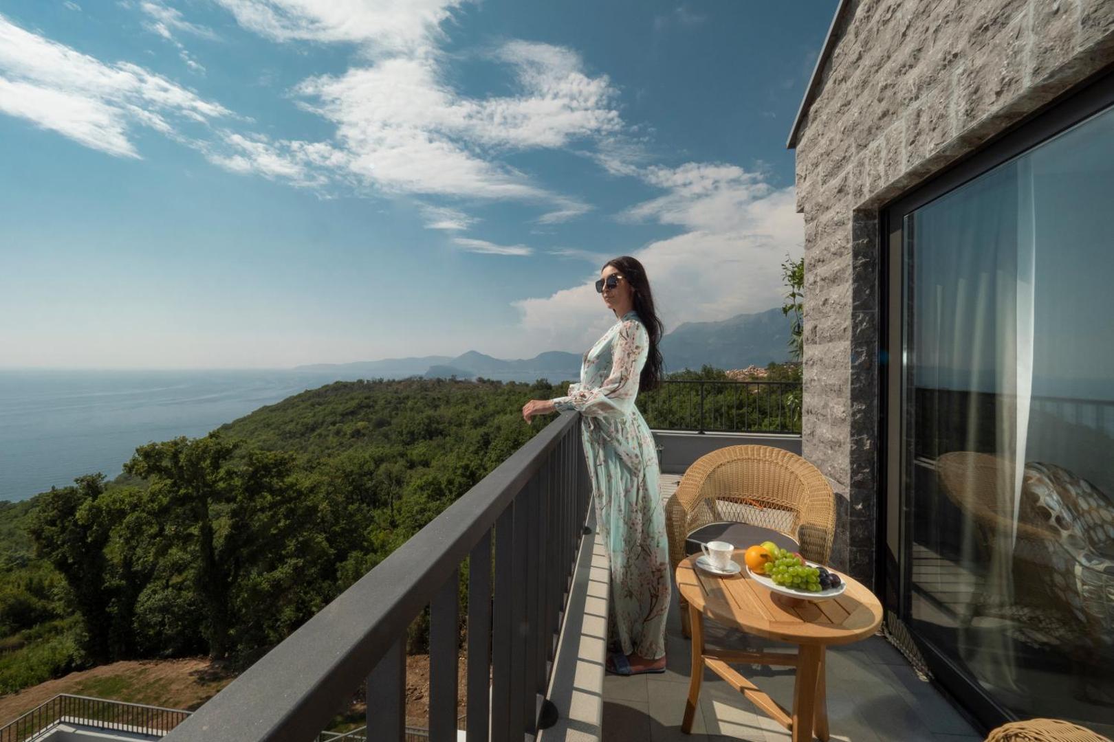 Vivid Blue Serenity Hotel numura balkons ar skatu uz Adrijas jūru un kalniem Melnkalnē — sieviete pie margām, galds ar augļiem