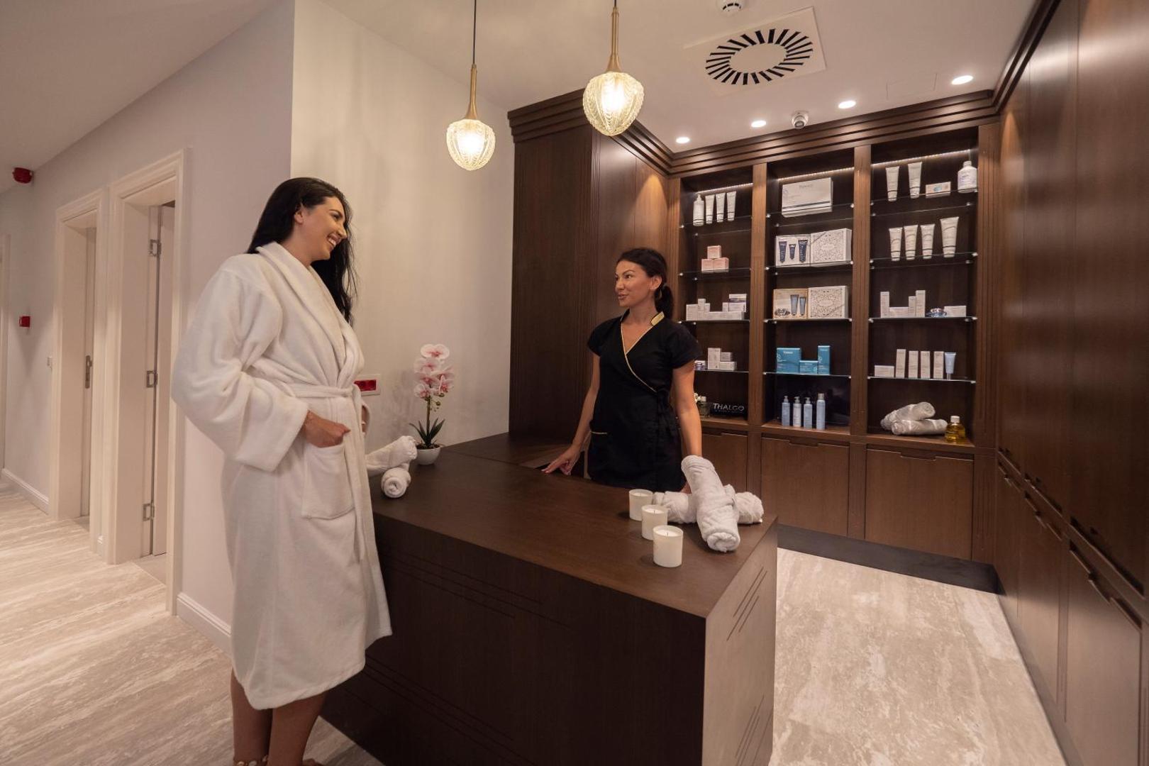 Vivid Blue Serenity Hotel spa centra recepcija Melnkalnē — viesis baltā halātā sarunājas ar darbinieci pie tumša koka letes