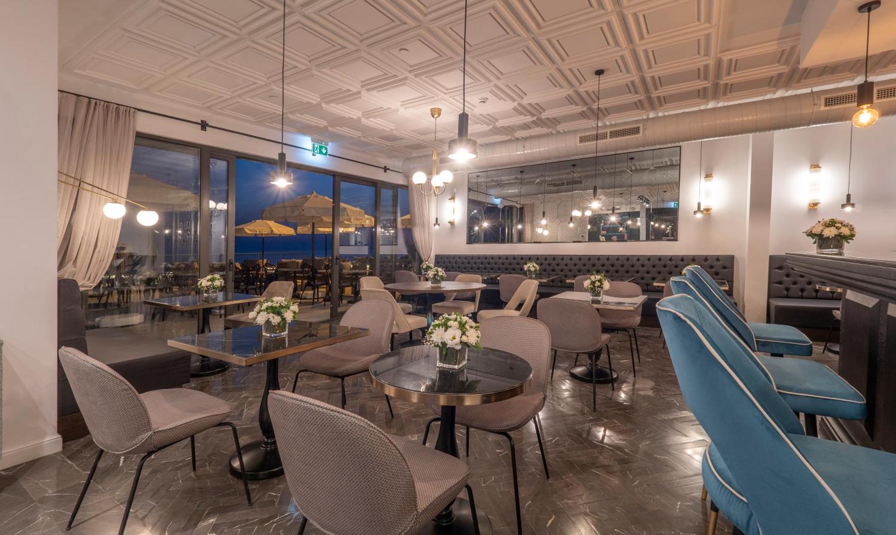 Vivid Blue Serenity Hotel restorāna iekštelpa vakarā ar elegantiem galdiņiem, ziliem bāra krēsliem un skatu uz terasi