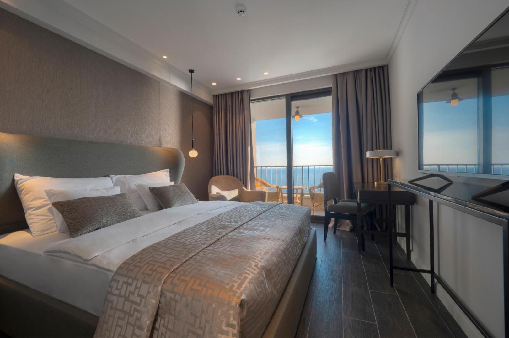 Mūsdienīgs Vivid Blue Serenity Hotel numura interjers ar tumša koka grīdu, ērtu gultu un panorāmas logu uz piekrasti
