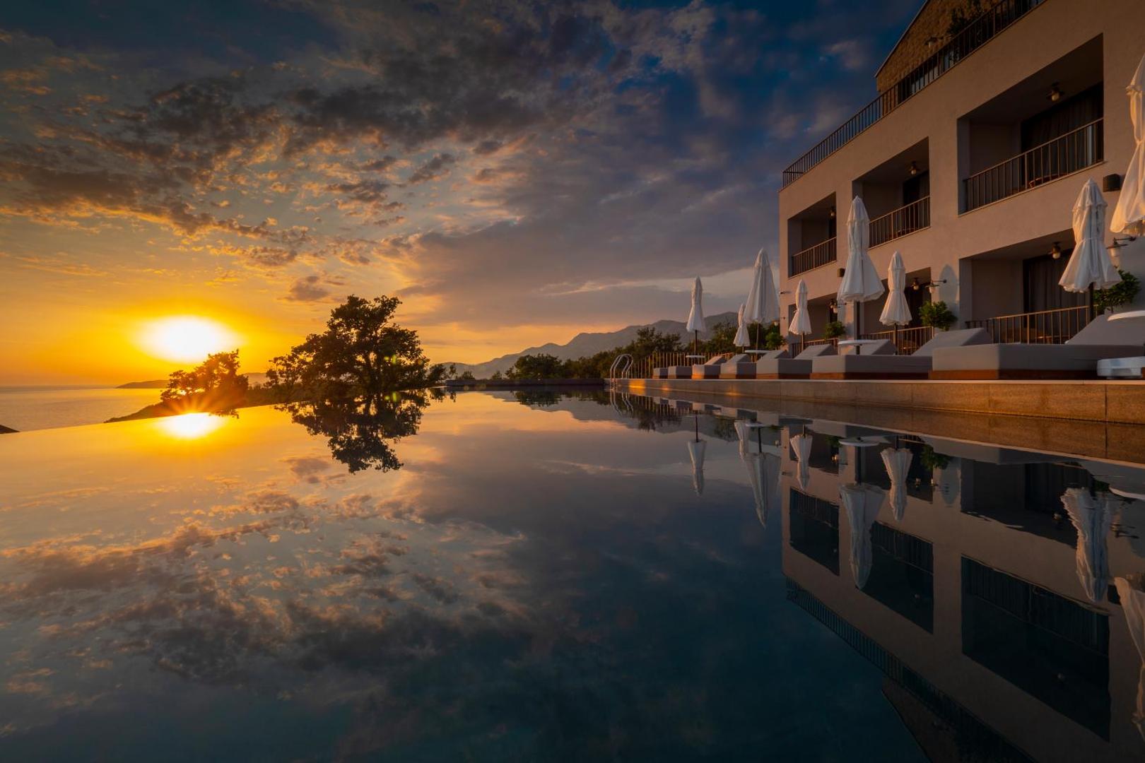 Vivid Blue Serenity Hotel ēkas fasāde un bezmalas baseins saulrietā — sauļošanās krēsli ar saulessargiem un kalnu silueti fonā