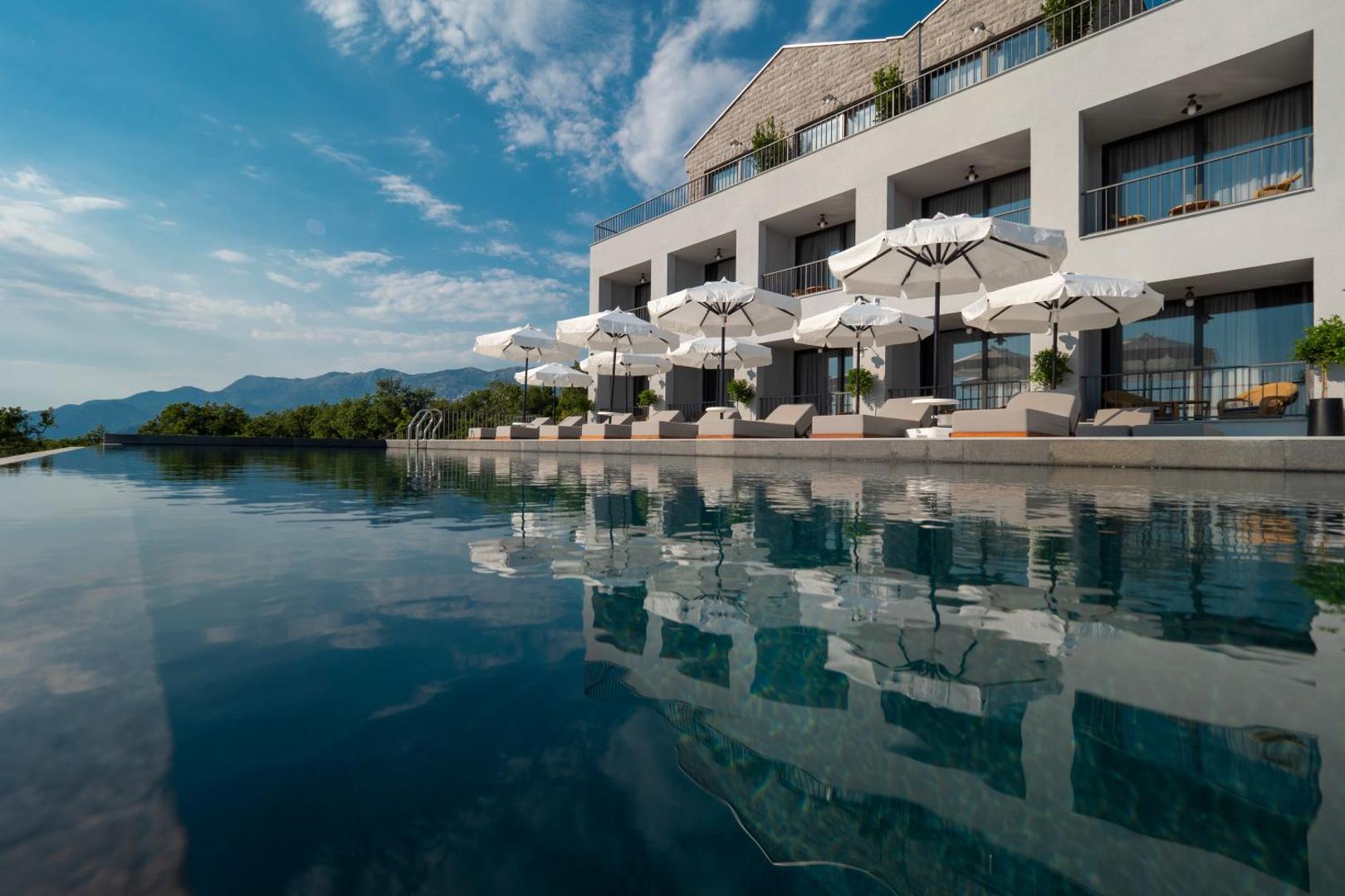 Vivid Blue Serenity Hotel āra bezmalas baseins ar sauļošanās krēsliem un saulessargiem Budvas rivjērā, Melnkalnē — skats uz viesnīcas fasādi
