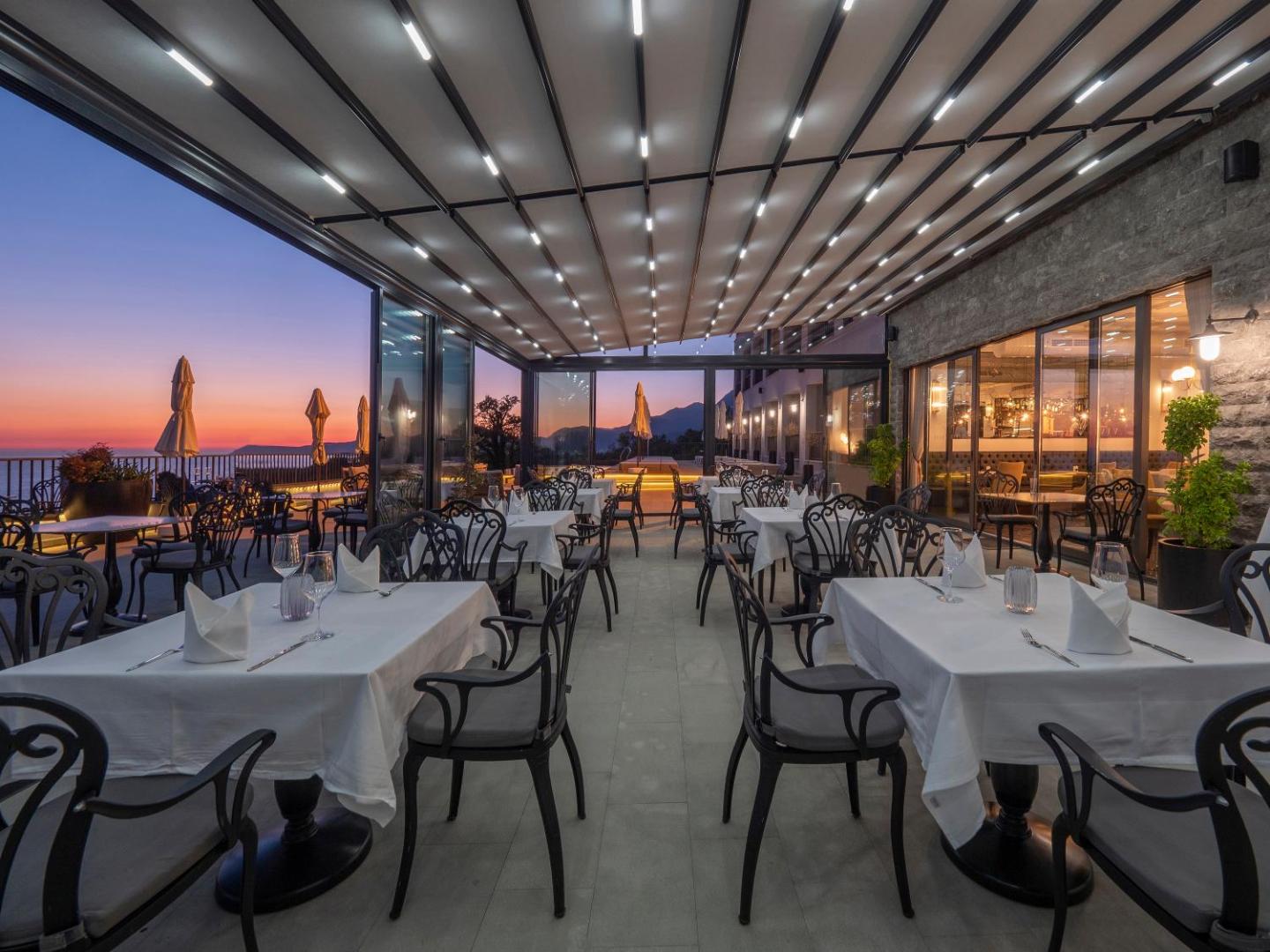 Vivid Blue Serenity Hotel restorāna āra terase ar klātiem galdiem un modernu apgaismojumu saulrieta laikā Melnkalnē