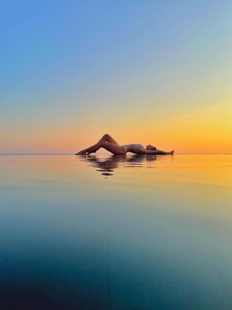 Vivid Blue Serenity Hotel bezmalas baseins saulrieta laikā — sieviete guļ ūdens virsmā, atspoguļojoties oranžajās debesīs