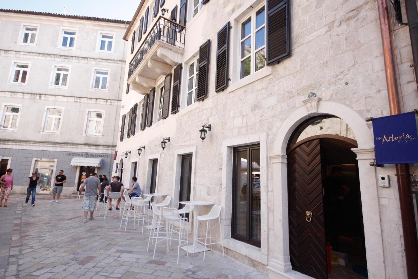 Astoria Boutique Hotel vēsturiskā akmens fasāde Kotorā ar āra terasi, baltiem krēsliem un koka slēģiem pie logiem
