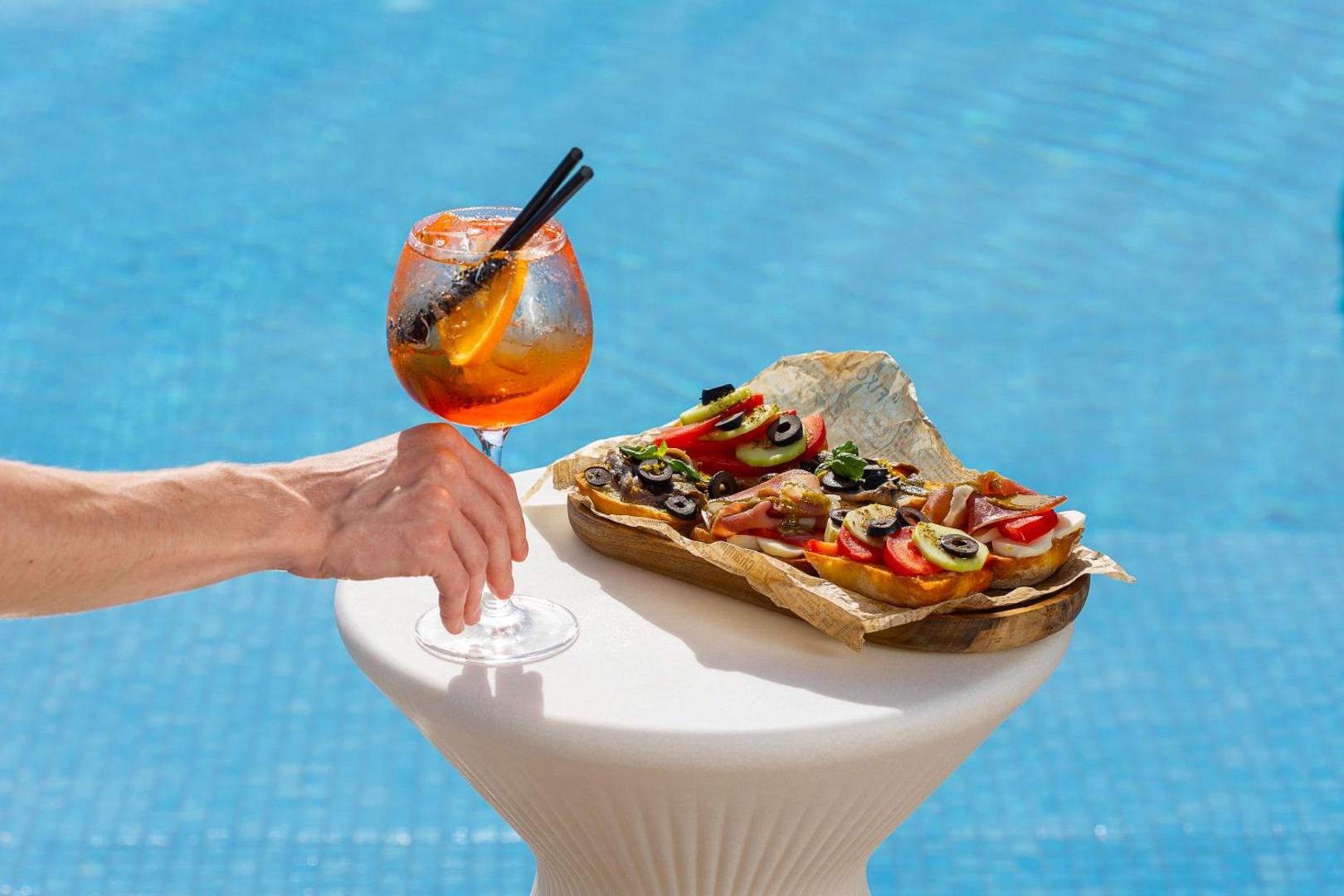 Aperol Spritz kokteilis un itāļu brusketas uz galdiņa pie Infinity by Dukley zilā peldbaseina