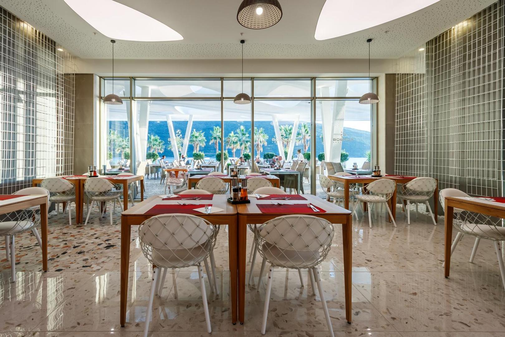 Carine Hotel Kumbor Superior restorāns ar modernām mēbelēm un lieliem panorāmas logiem, no kuriem paveras skats uz palmu dārzu un jūru.
