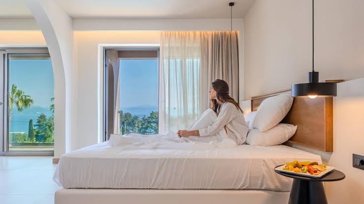 Sieviete atpūšas Iolida Corfu Resort & Spa gultā ar skatu uz dārzu un jūru caur lieliem logiem