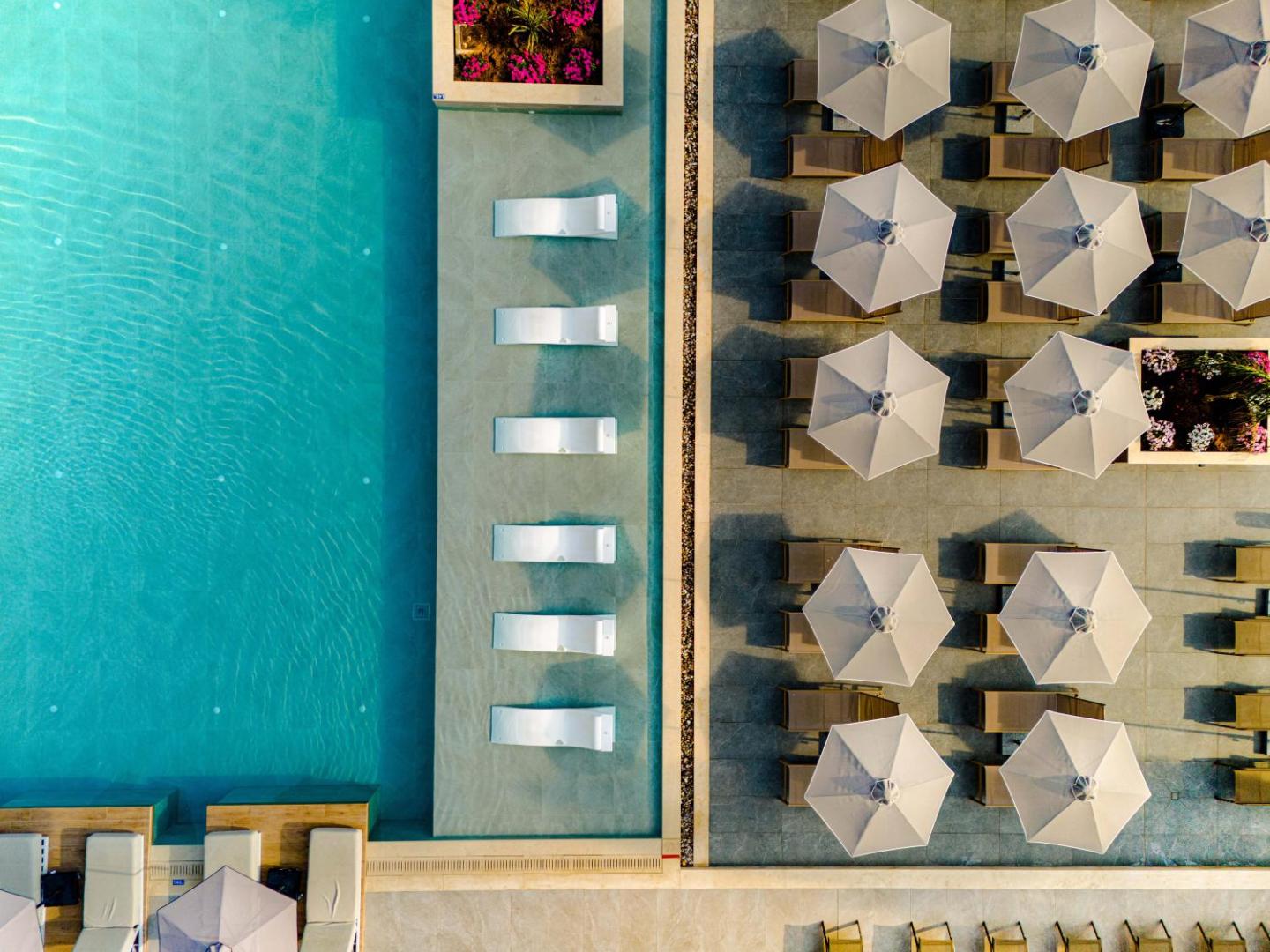 Iolida Corfu Resort & Spa āra baseins no putna lidojuma — tirkīza ūdens, balti saulessargi un gultas seklajā zonā