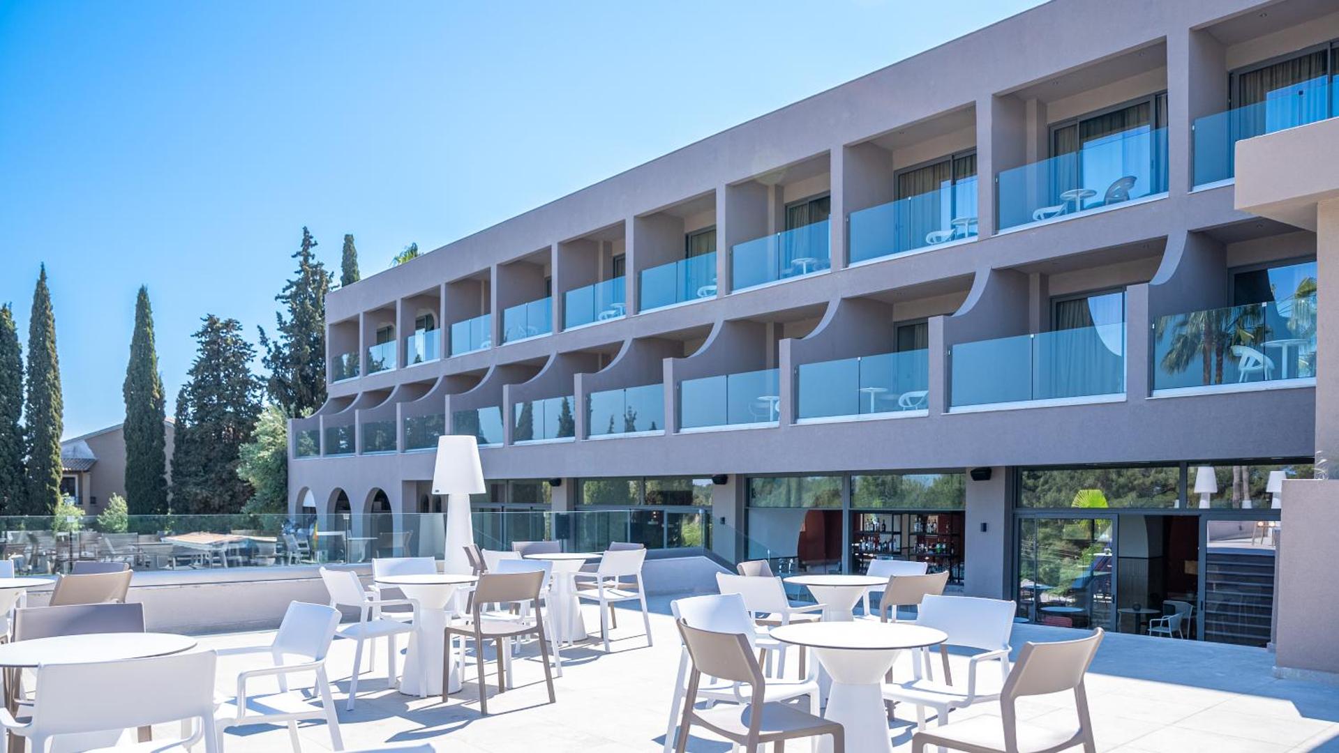 Iolida Corfu Resort & Spa āra terase ar baltiem galdiem un krēsliem, fonā redzami numuri ar stikla balkoniem