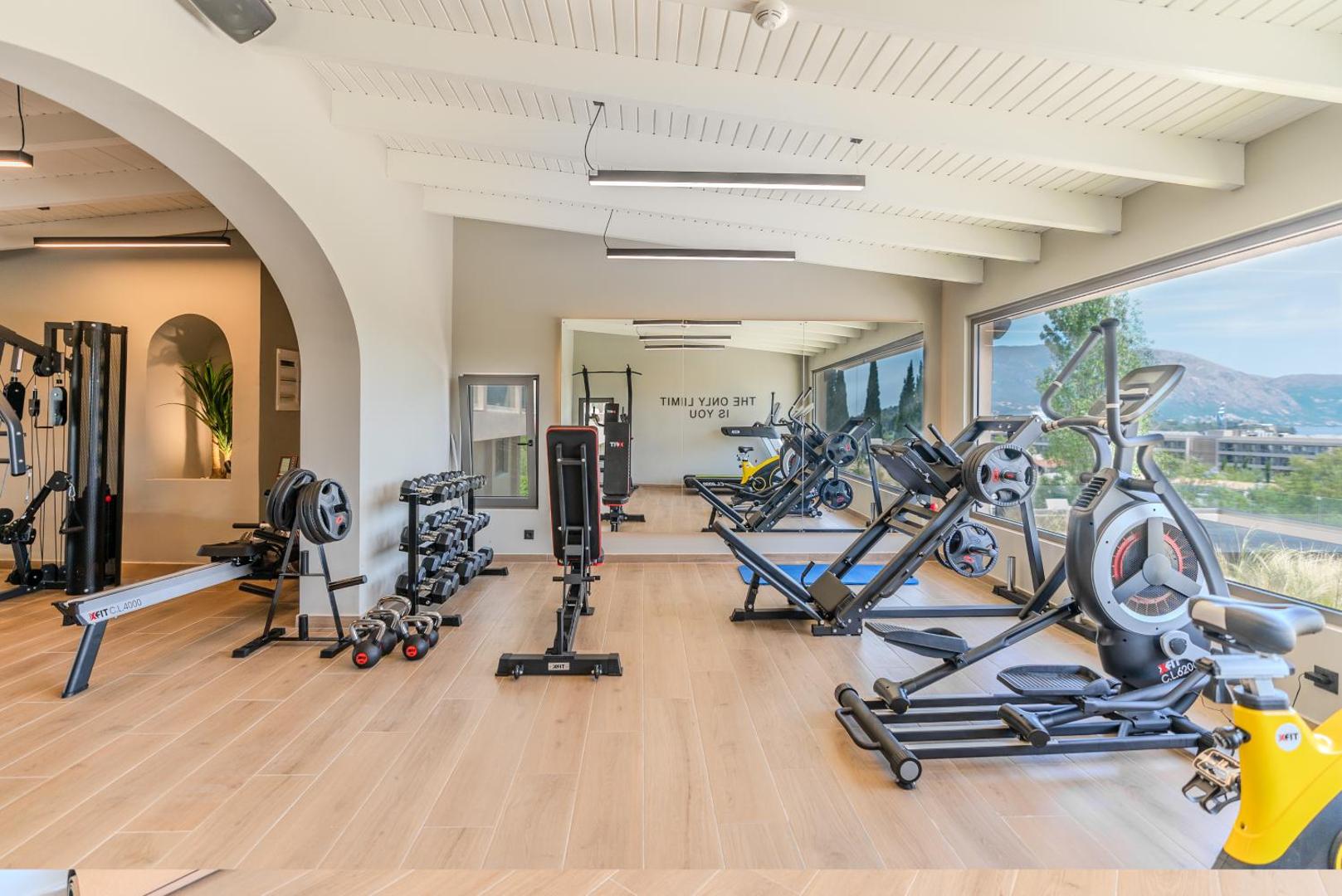 Iolida Corfu Resort & Spa fitnesa centrs ar moderniem trenažieriem un skatu uz kalniem caur lieliem logiem