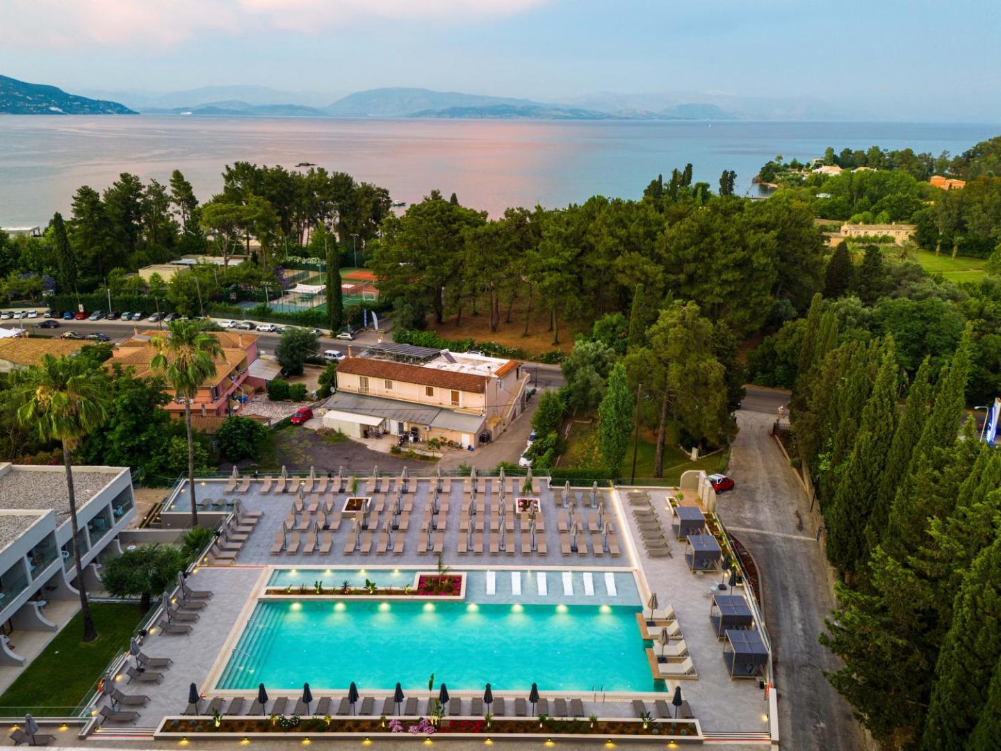 Iolida Corfu Resort & Spa panorāmas skats uz Jonijas jūru, piekrastes priežu mežu un viesnīcas baseina zonu saulrietā