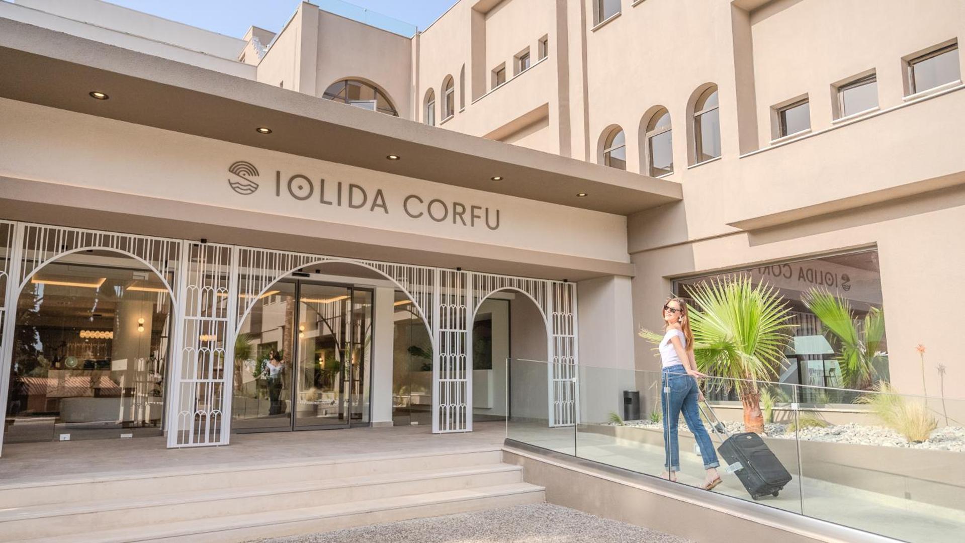 Viesis ar koferi pie Iolida Corfu Resort & Spa ieejas — moderna viesnīcas ēka ar gaišu fasādi un palmām Korfu