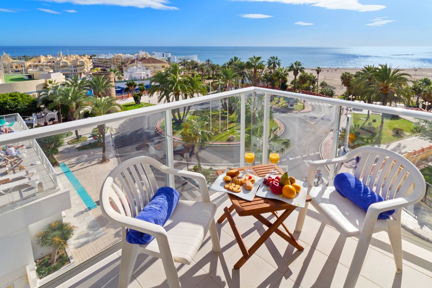Balkons ar skatu uz jūru, kur ir divas baltas krēsli un koka galdiņš ar brokastu izlasi, tostarp augļiem un dzērieniem. Apkārt ir palmām un pludmale. panoramic view with beach and pools