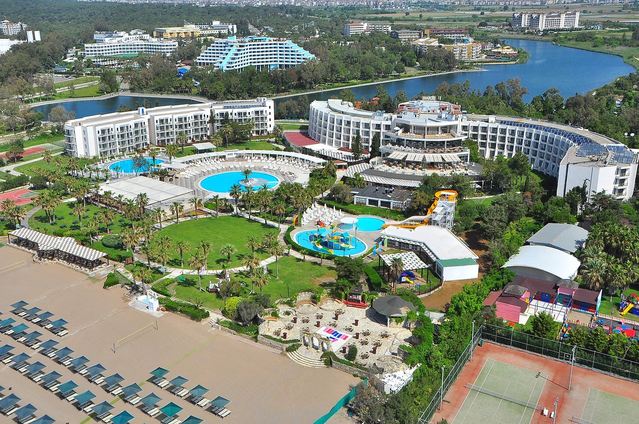 Liela kūrorta zona ar peldbaseiniem, slidkalniņiem un zaļām ainavām, kas atrodas pie ūdens. Redzamas dažādas atpūtas zonas un sporta laukumi. panoramic view with beach and pools