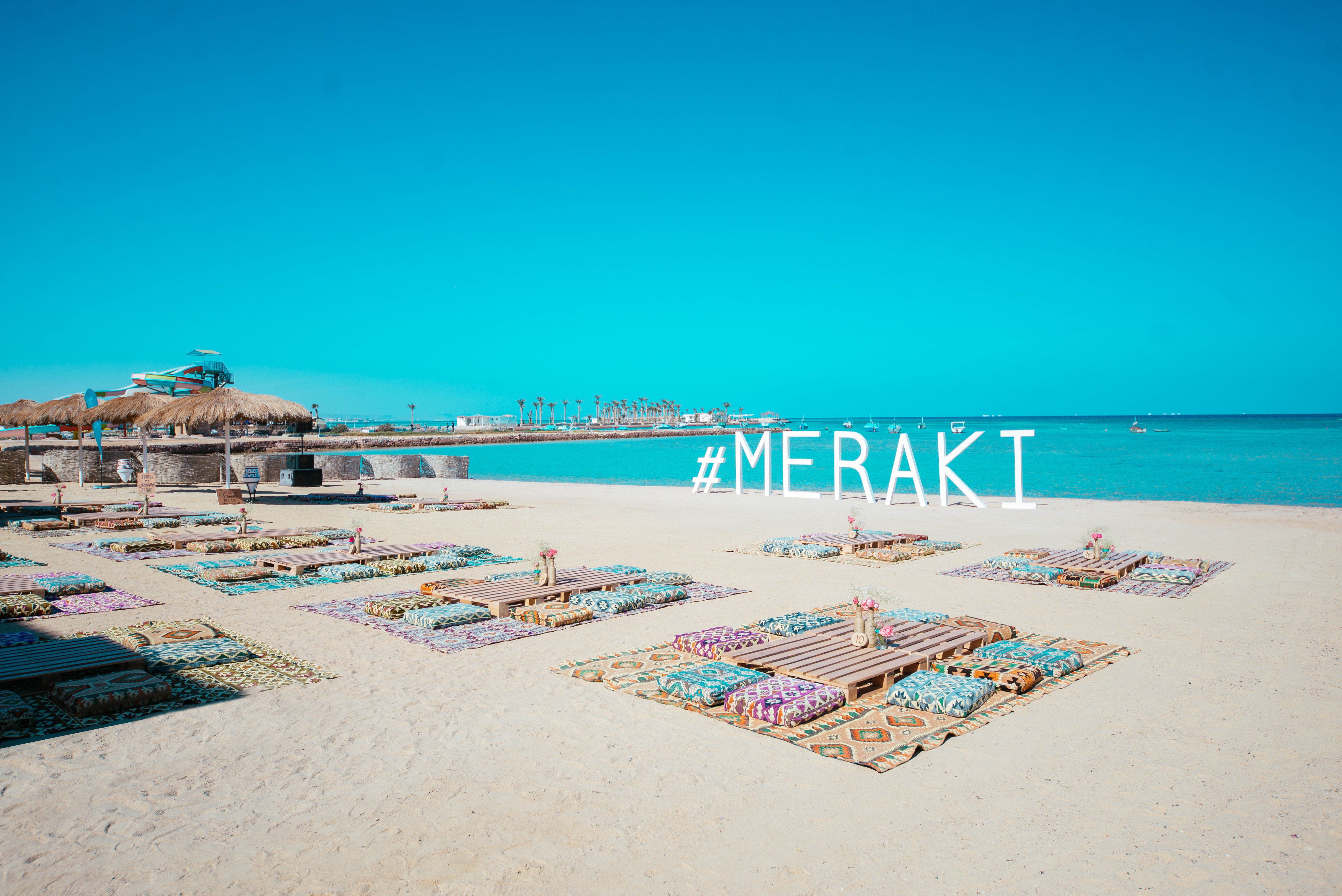 Pludmale ar krāsainām paklājām un dekoratīvām puķēm, kur centrālais elements ir liels uzraksts "#MERAKI". Jūras fons ar skaidru zilu debesi un palmām. panoramic view with beach and pools