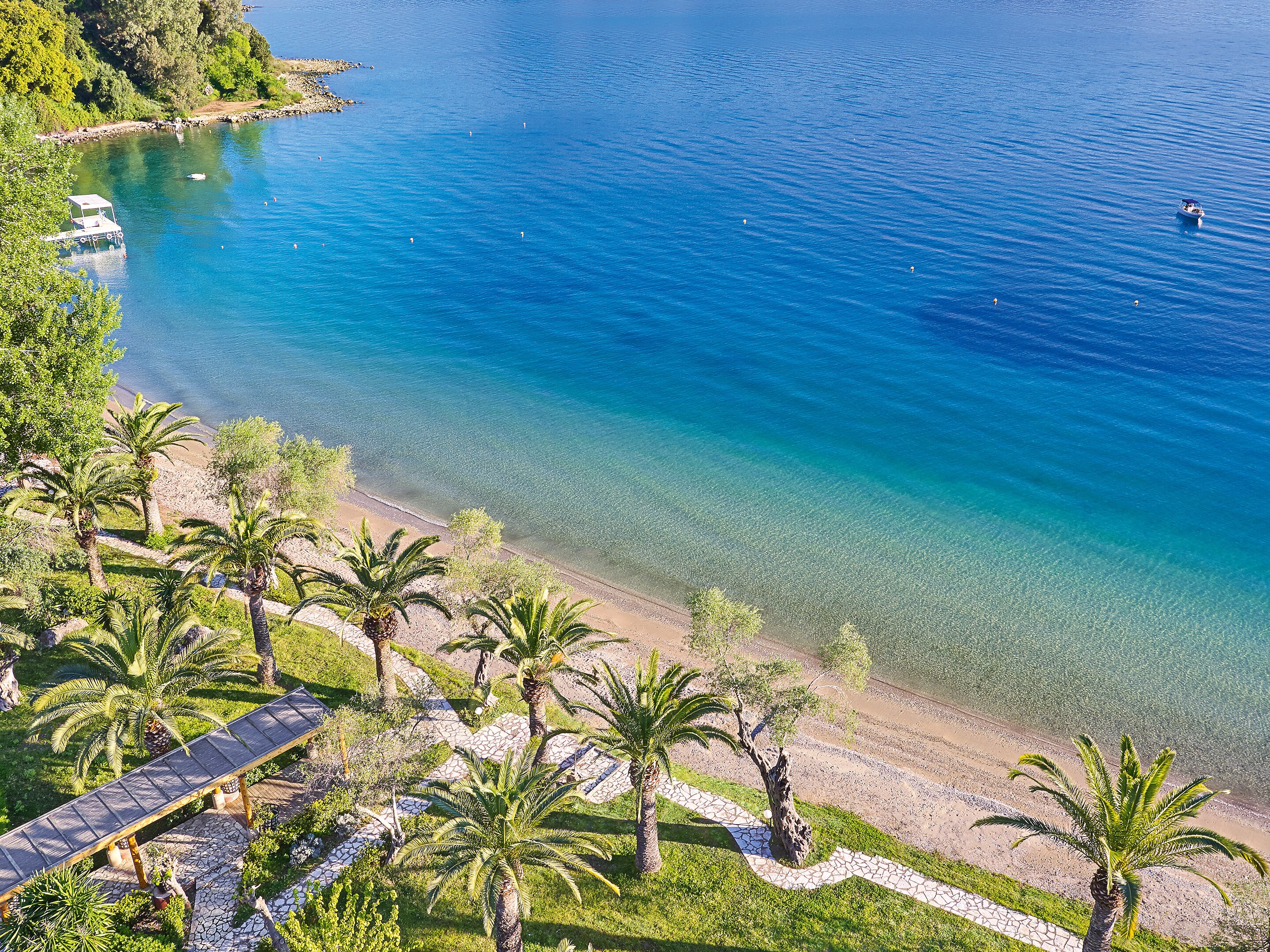 Skaista pludmale ar palmām un skaidru zilo ūdeni, kas mierīgi viļņojas. Fons ietver krūmus un laivu, radot relaksējošu atmosfēru. panoramic view with beach and pools