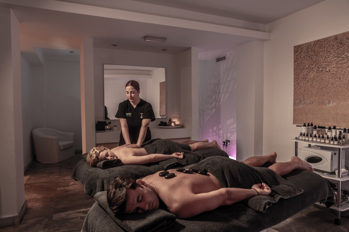 Relaksējoša masāža spa centrā, kur masieris apkalpo divus klientus, kuri guļ uz masāžas galdiem, pārklāti ar dvieļiem. Sienās redzami dekoratīvi elementi un aromātiskas eļļas.