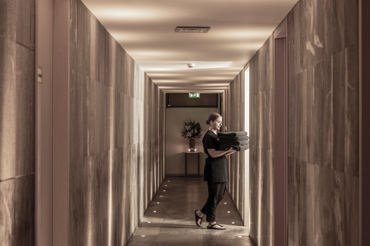 Skaists spa koridors ar darbinieku, kurš nes vairākus dvieļus. Telpa ir apgaismota ar mīkstu gaismu, radot relaksējošu atmosfēru.