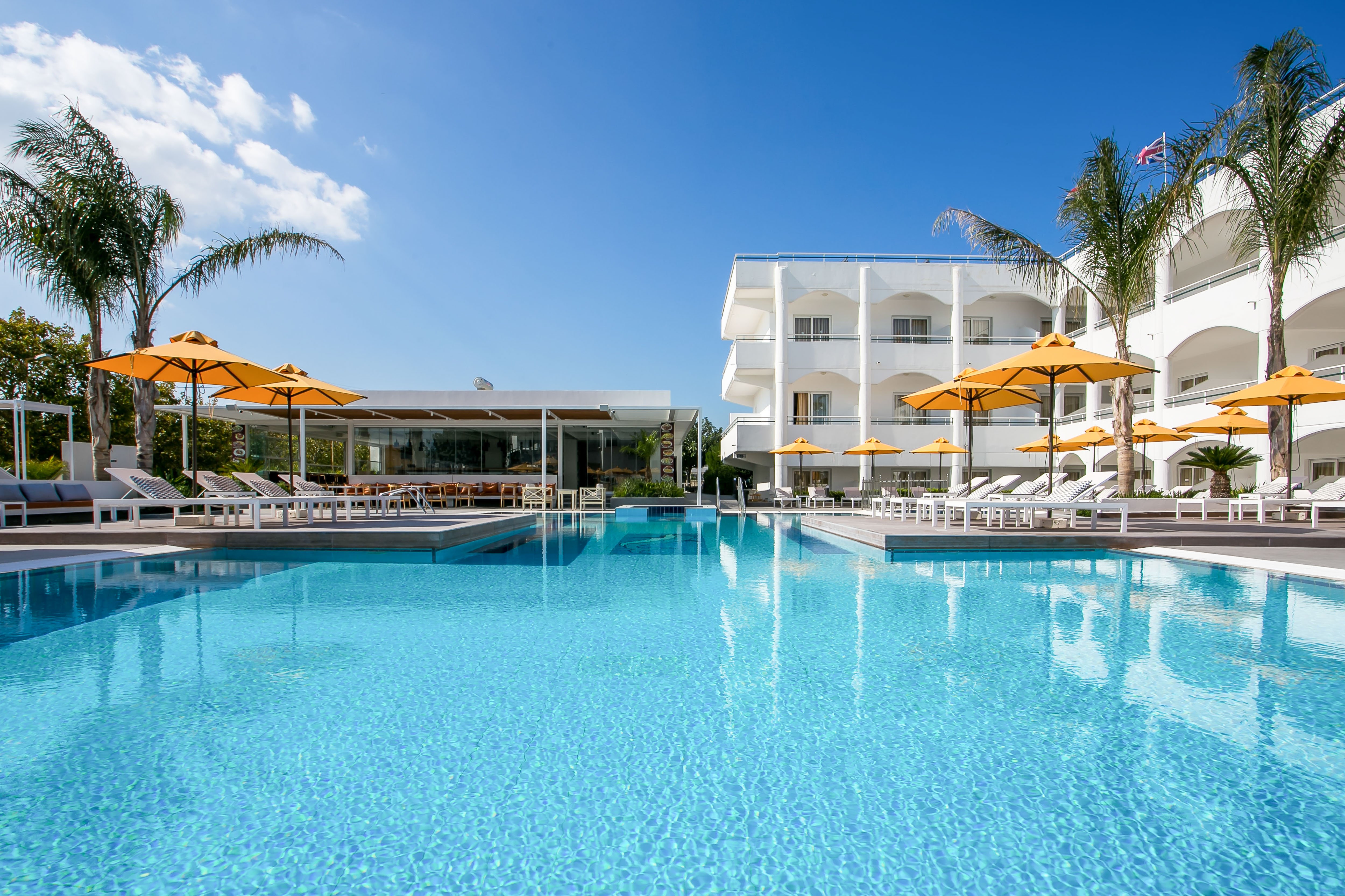 Peldbaseins ar skaidru zilu ūdeni un oranžiem saulessargiem, blakus modernai viesnīcai. Ideāla vieta atpūtai un relaksācijai. panoramic view with beach and pools