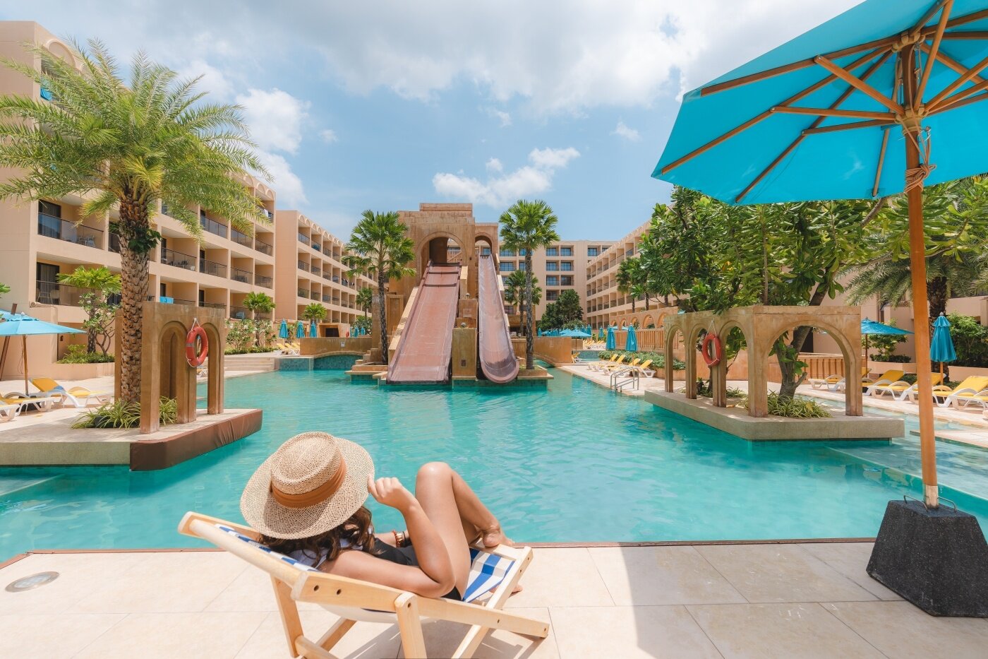 Vasaras brīvdienu kūrorts ar peldbaseinu un slidkalniņu, kurā cilvēks atpūšas uz krēsla zem saulessarga. Apkārt palmām un viesnīcu ēkām ir daudz saules krēslu.