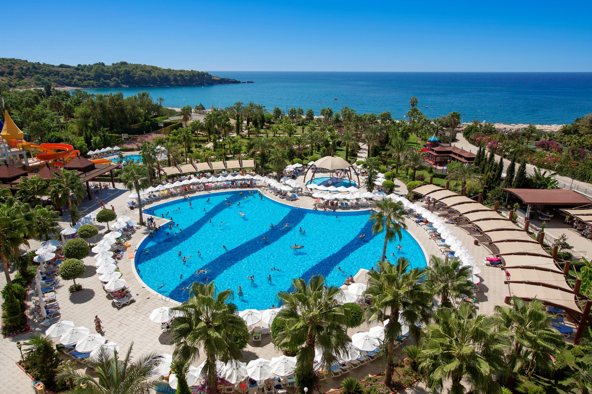 Viesnīcas baseins ar zilu ūdeni un saulessargiem, apkārt palmām un ziedošām augu kompozīcijām. Fons veido skaists skats uz jūru un kalniem. panoramic view with beach and pools
