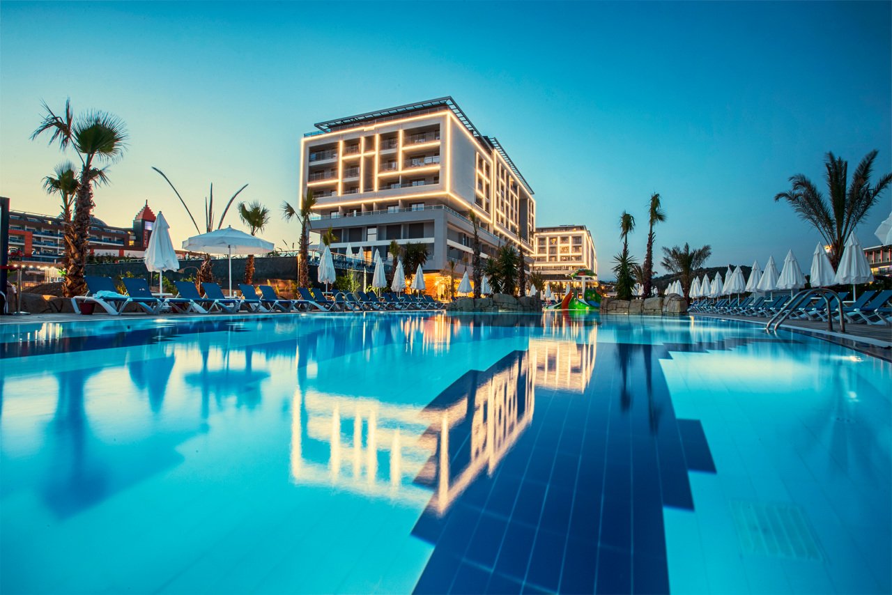 Mūsdienīgs viesnīcas komplekss pie baseina ar palmām un sauļošanās krēsliem, kas atspoguļojas ūdenī, radot relaksējošu atmosfēru. panoramic view with beach and pools