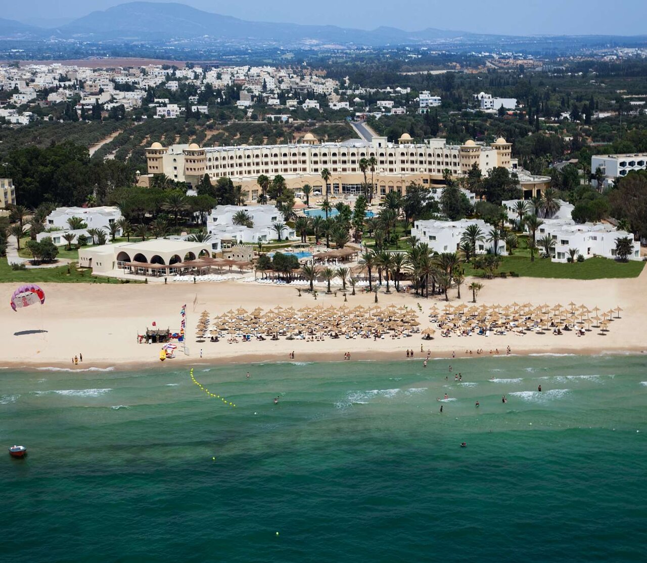 Plašs skats uz pludmali ar saulessargiem, kūrortu un zaļajiem kokiem fona. Jūras viļņi krastā un cilvēki izklaidējas ūdenī. panoramic view with beach and pools